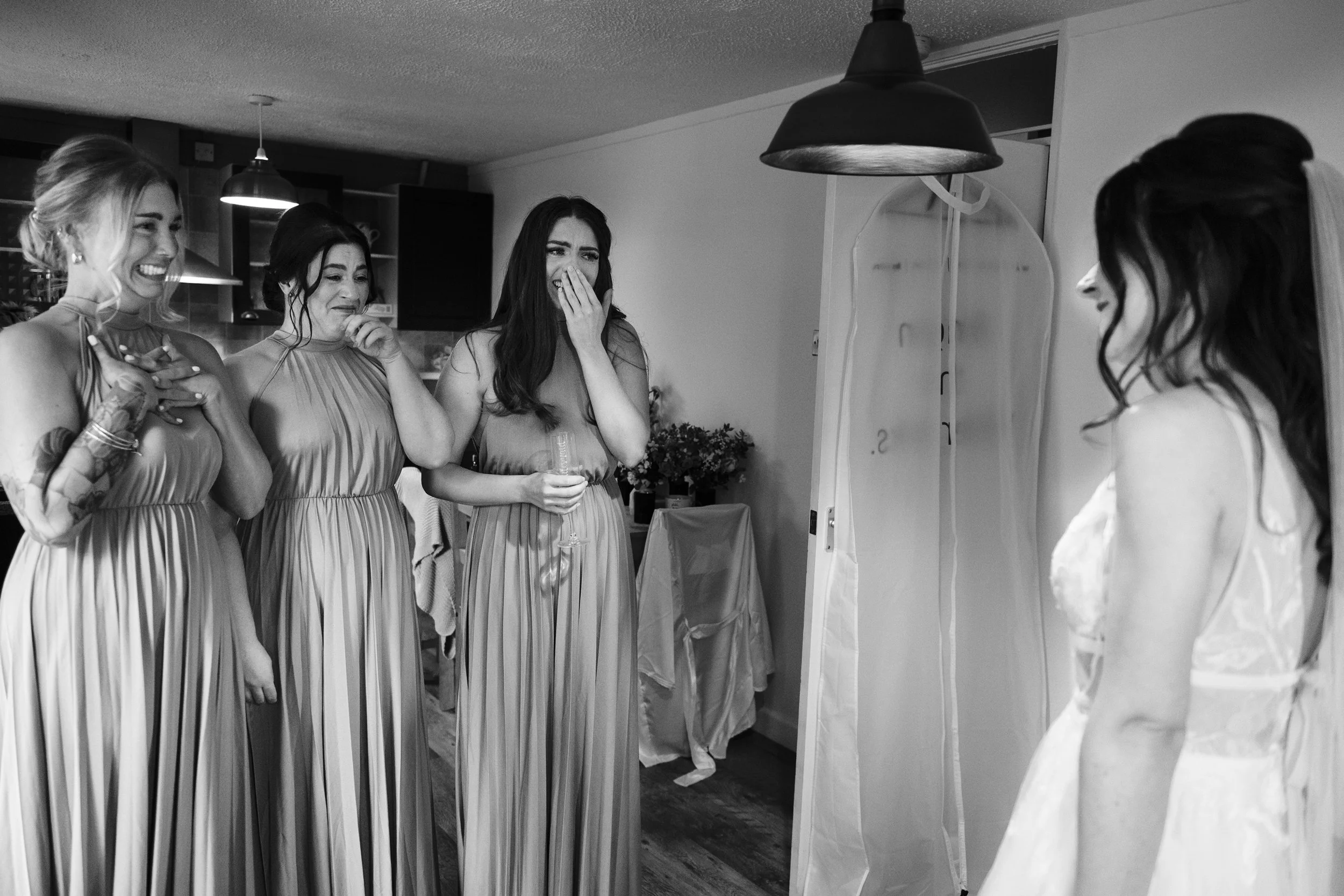 Four women in dresses standing in front of a bride, who is on the right, at a wedding preparation.