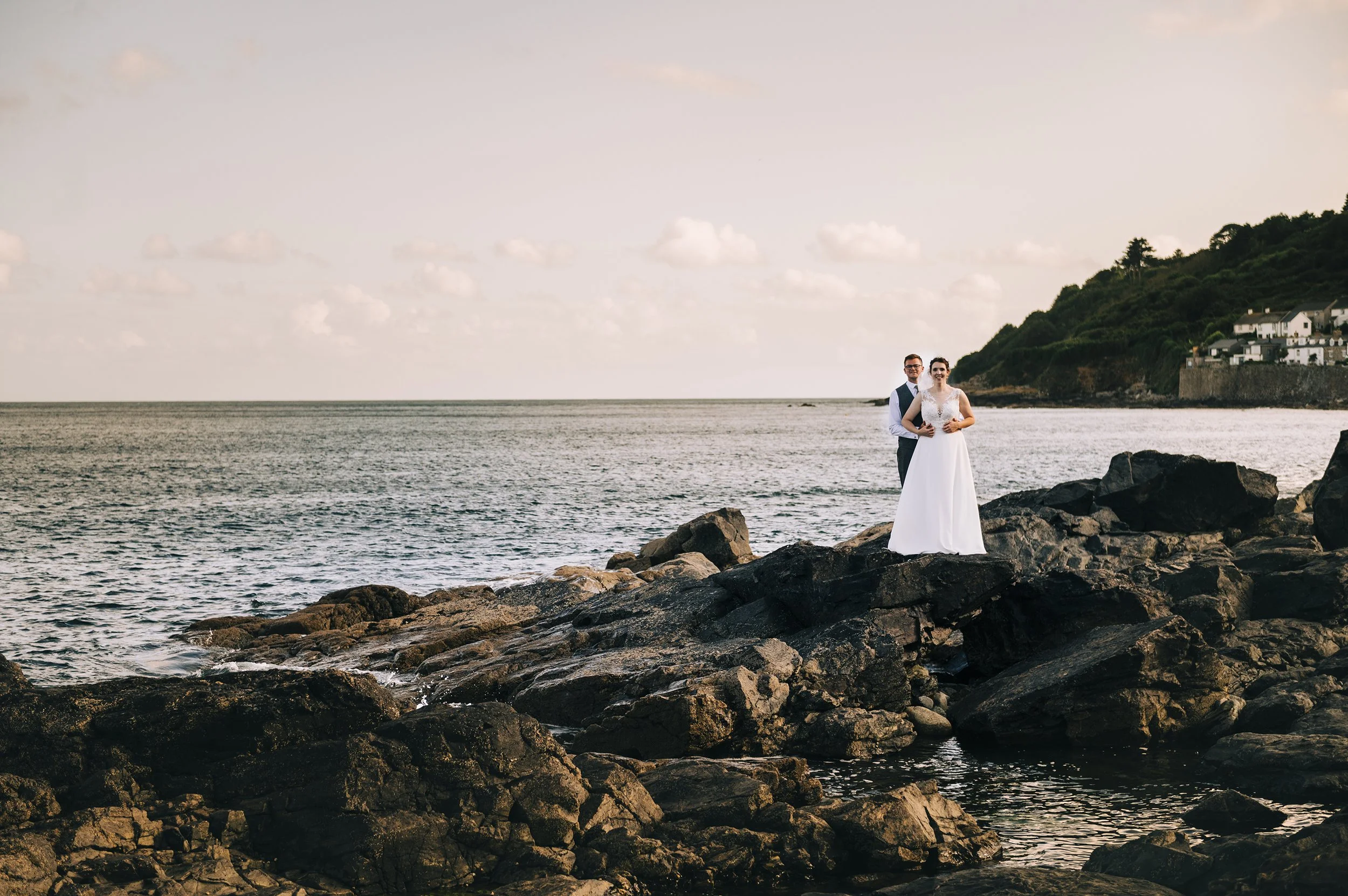 A bride and groom standing on a rocky shoreline with the ocean and a distant hillside with houses in the background.