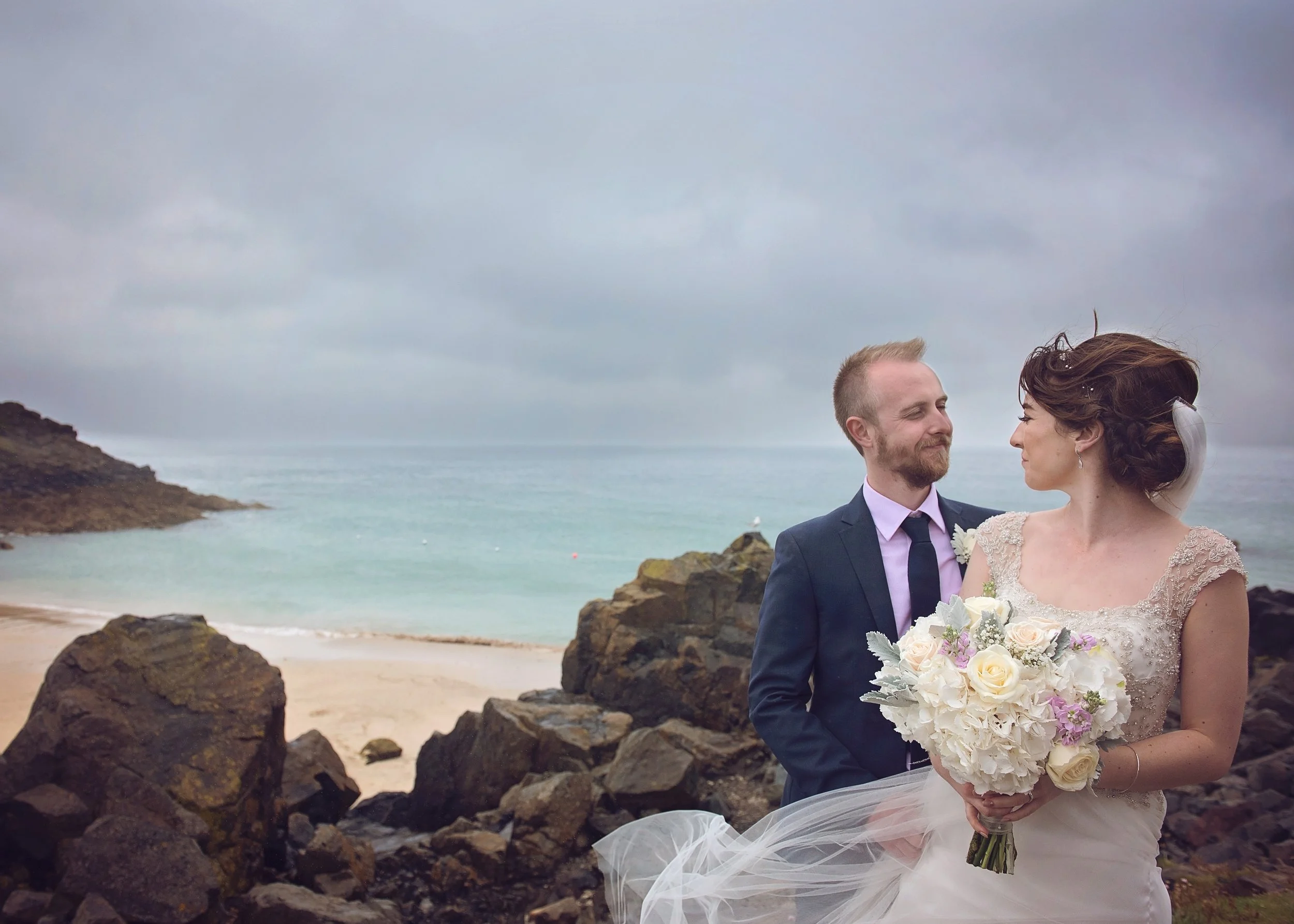 A bride and groom standing on rocks near a beach with an overcast sky, the bride holding a bouquet of white and purple flowers and the groom looking at her.