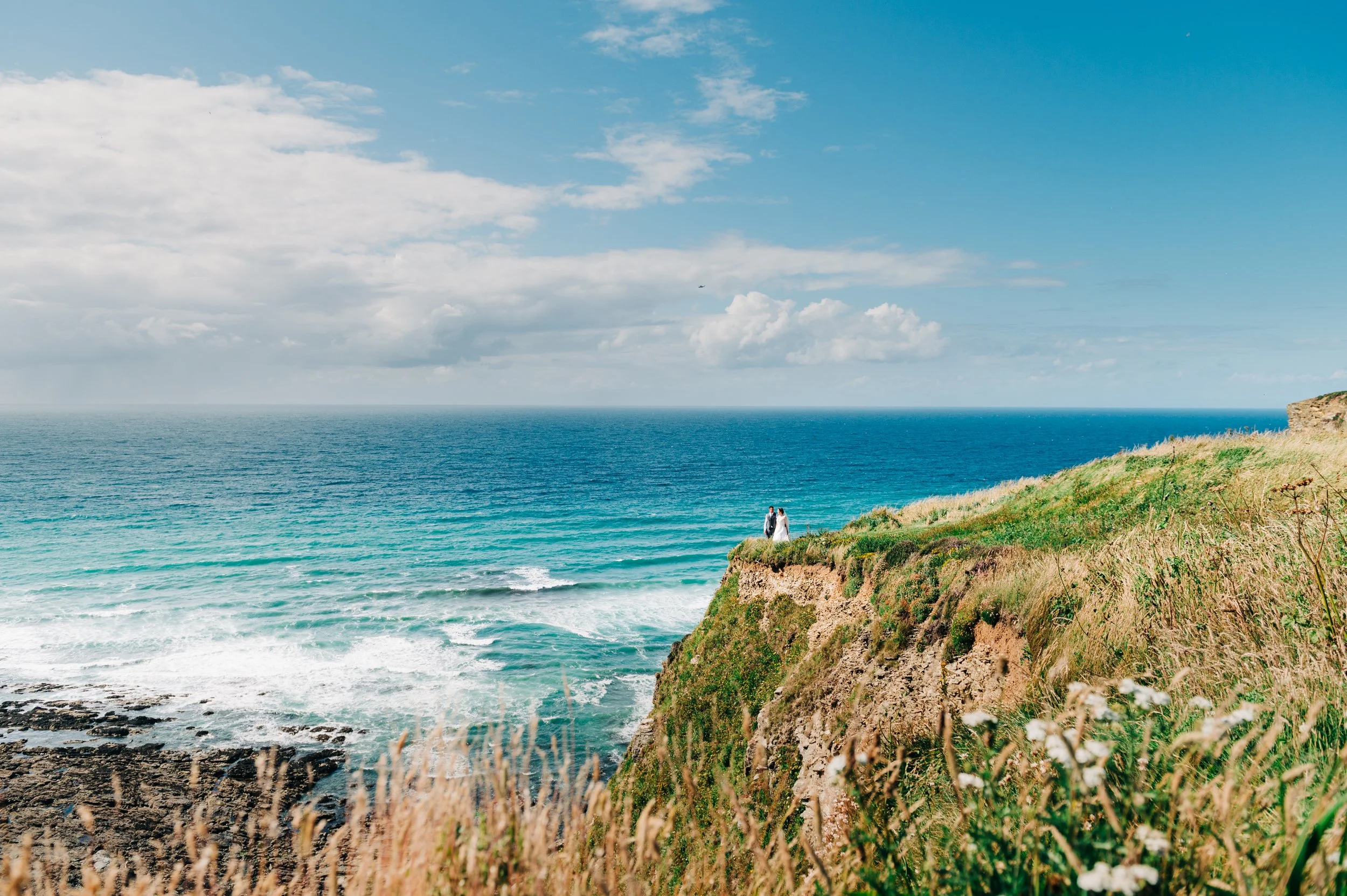 A couple dressed in wedding attire standing on a grassy cliff overlooking the ocean with blue sky and clouds in the background.