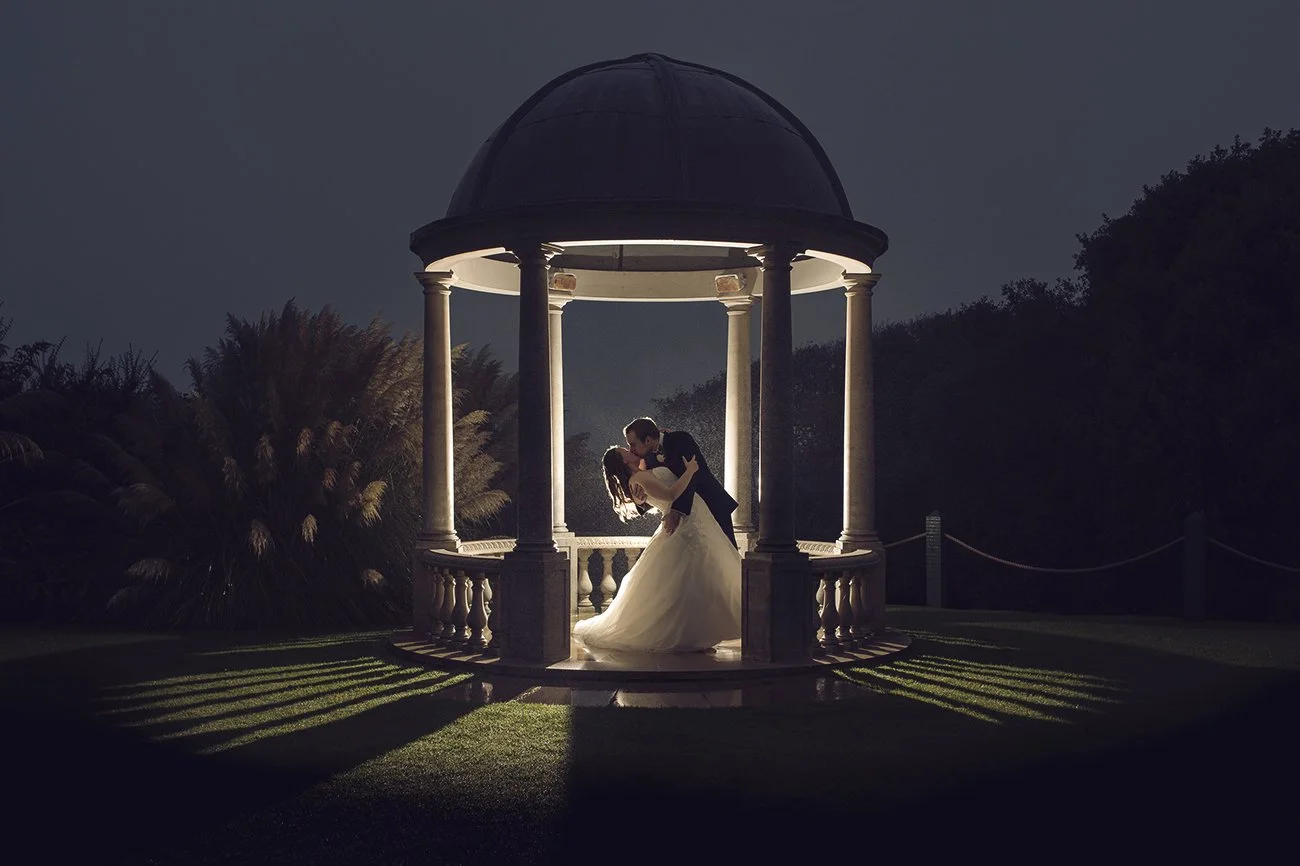 A bride and groom share a kiss under a lit gazebo at night, with shadows cast on the grass below.
