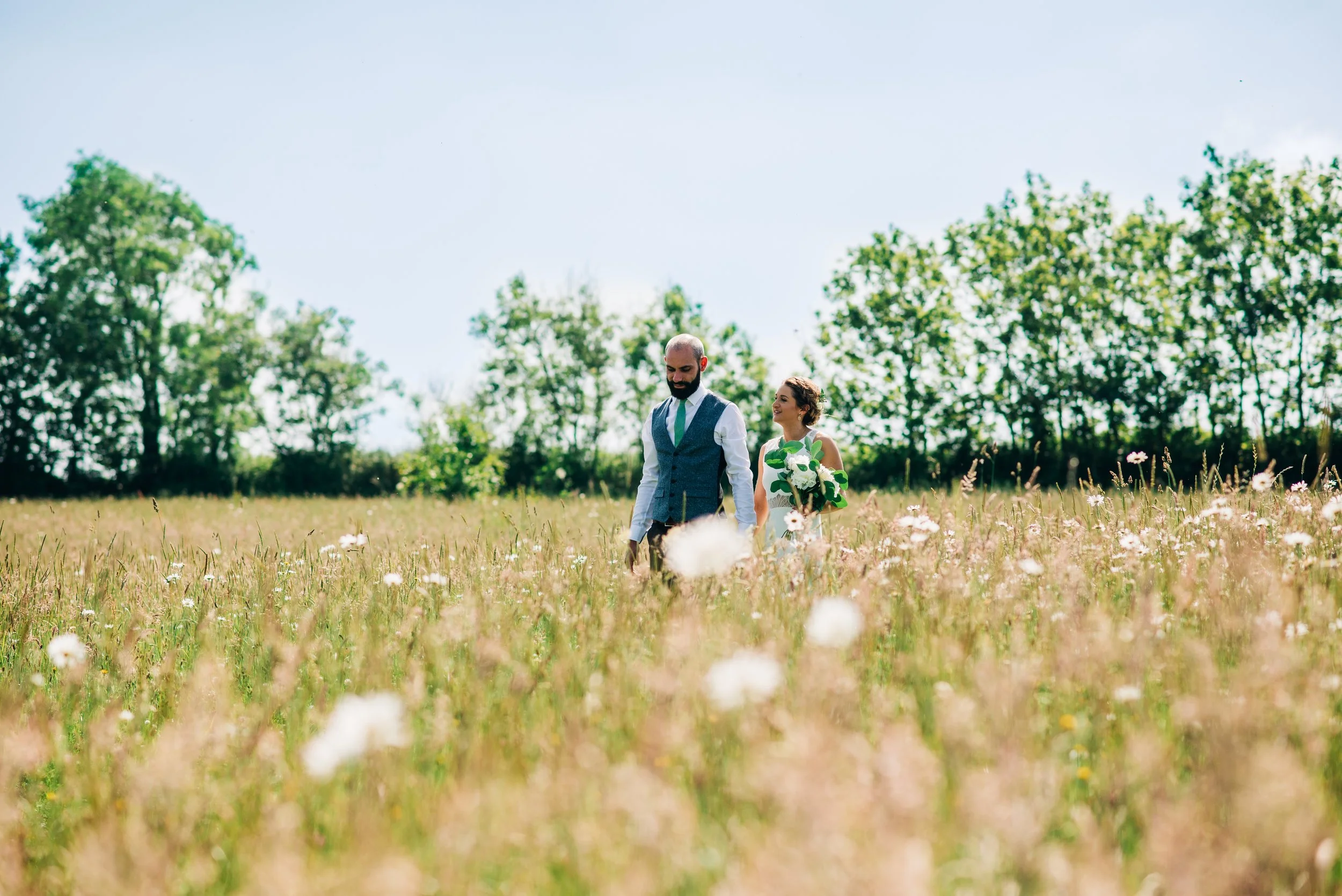A couple walks through a field of wildflowers with trees in the background on a sunny day, the woman holding a bouquet of flowers.