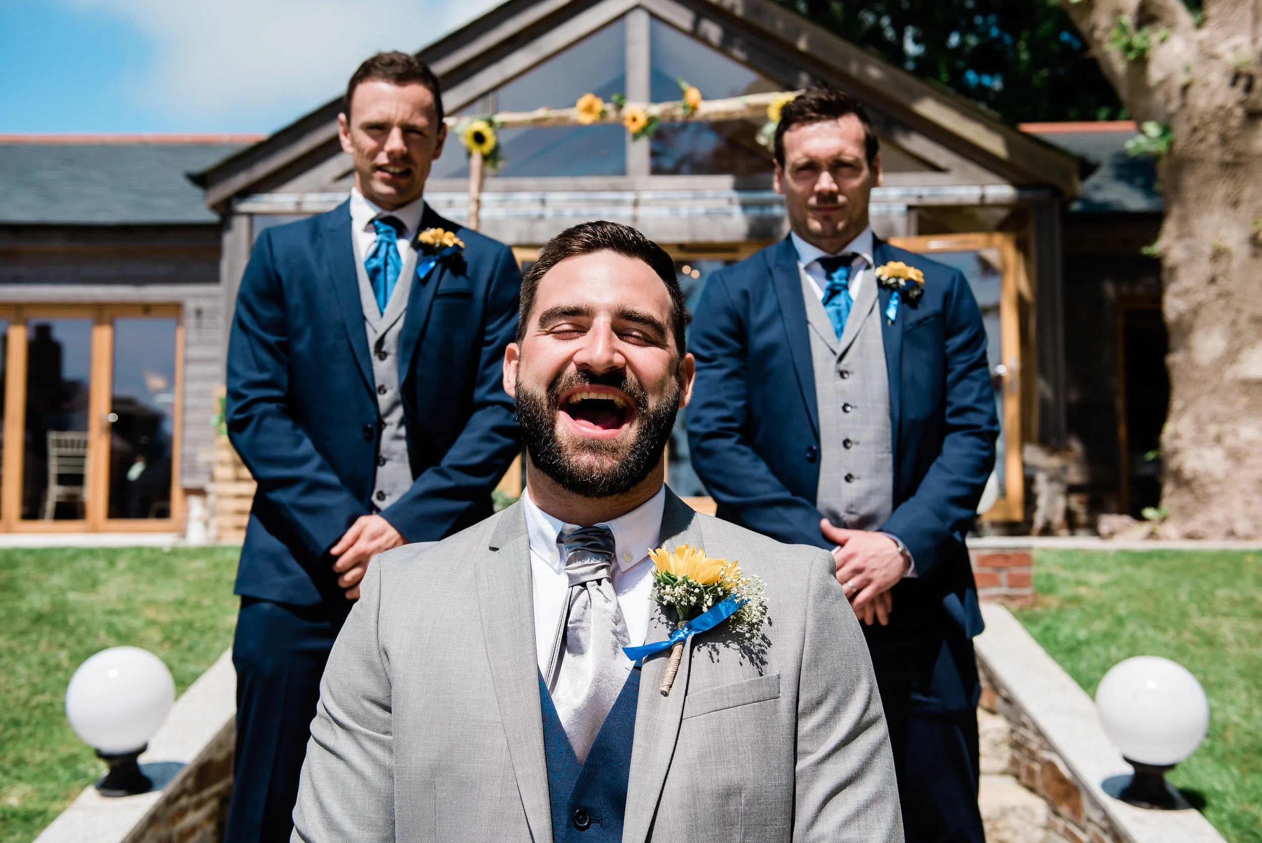 A smiling groom with a beard, wearing a gray suit and floral boutonniere, stands in front of three groomsmen in navy suits and blue ties outside a building with a glass roof and decorated with sunflowers, on a sunny day.