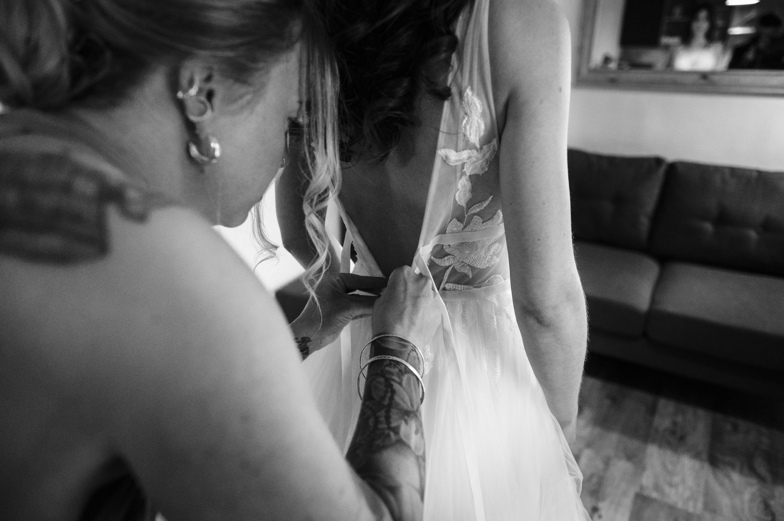 A person helping a bride zip up her wedding dress in a room with a couch and mirror.