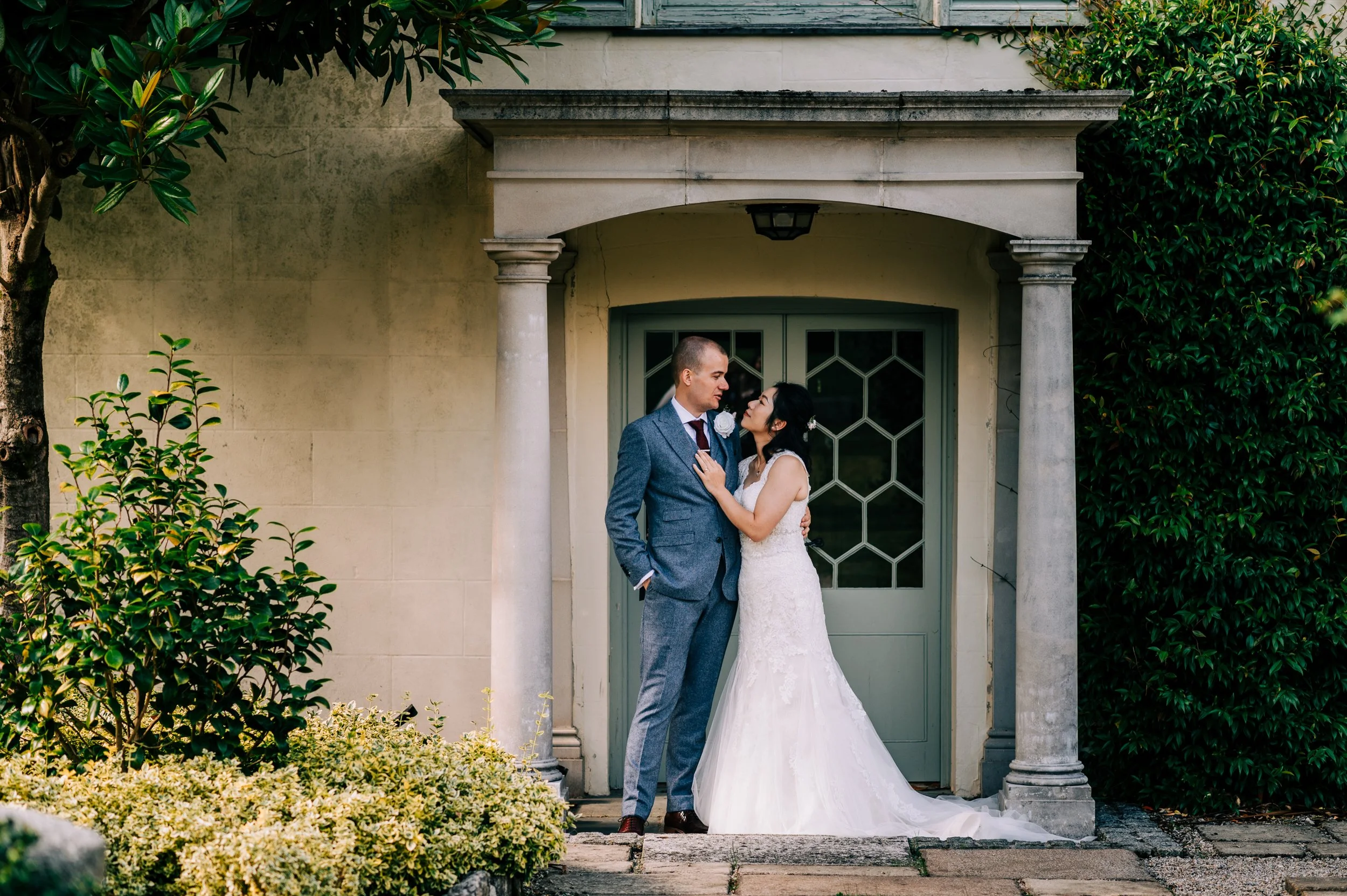 A newlywed couple stands close together in front of a vintage green door, on a porch with stone columns, surrounded by greenery. The groom is in a blue suit, and the bride is in a white lace wedding dress.