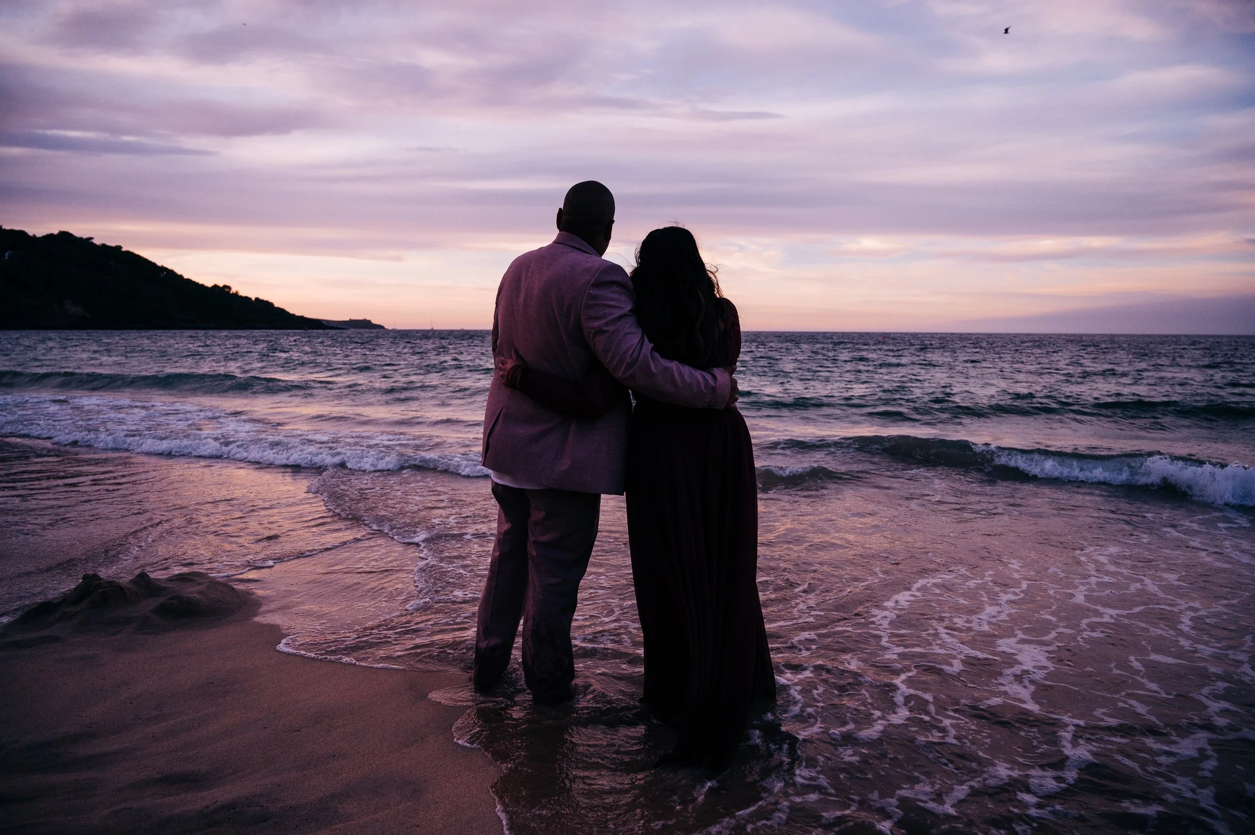 A couple standing on the beach at sunset, embracing and watching the ocean.