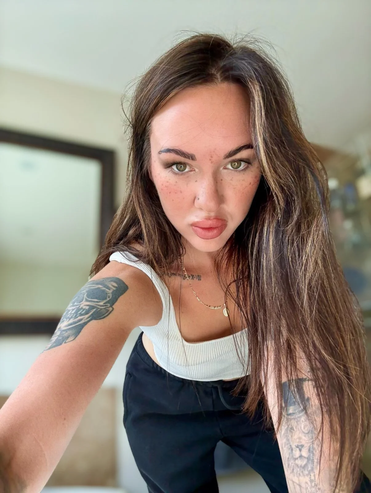 A young woman with long brown hair and tattoos on her arms is taking a selfie indoors, looking directly at the camera with a serious expression.