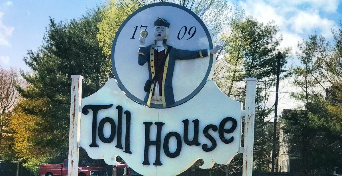 A white sign with black text reading 'Toll House' and a clock on top showing the time 17:09, topped with a figure of a colonial man in costume pointing and holding a mug, outdoors with trees in the background.