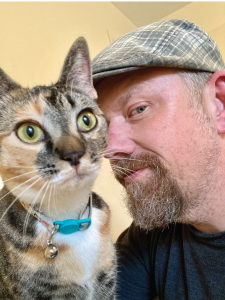 A man with a gray beanie and beard taking a selfie with a tabby cat with green eyes and a blue collar.