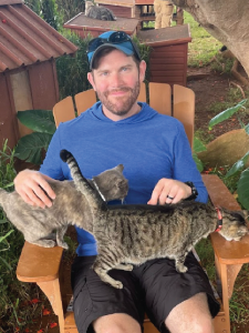 Man sitting outdoors in a blue shirt and baseball cap, holding two tabby cats, one on his lap and the other on the armrest, with trees and gardening tools in the background.
