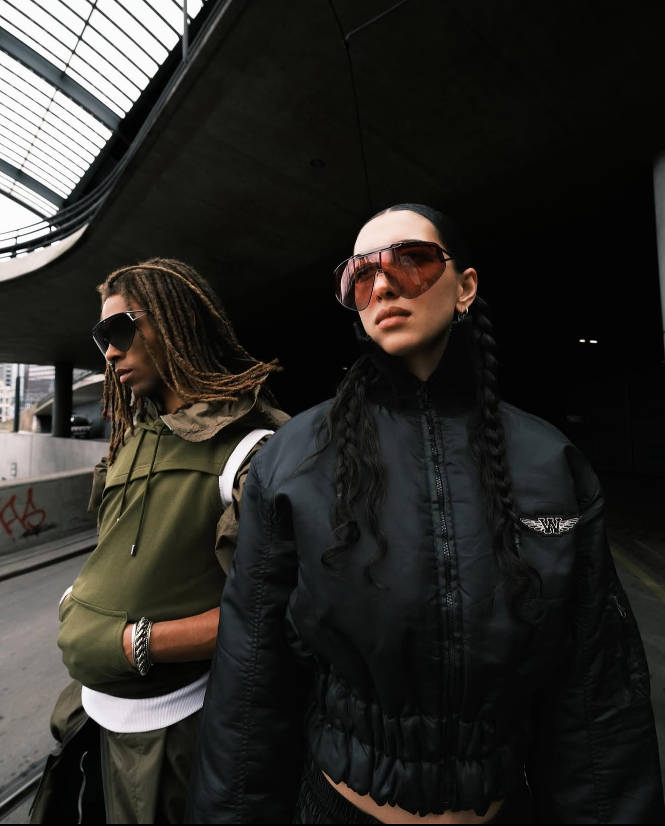 Two women with sunglasses standing under an urban overpass, one with dreadlocks and green hoodie, the other with long braided hair and black jacket.