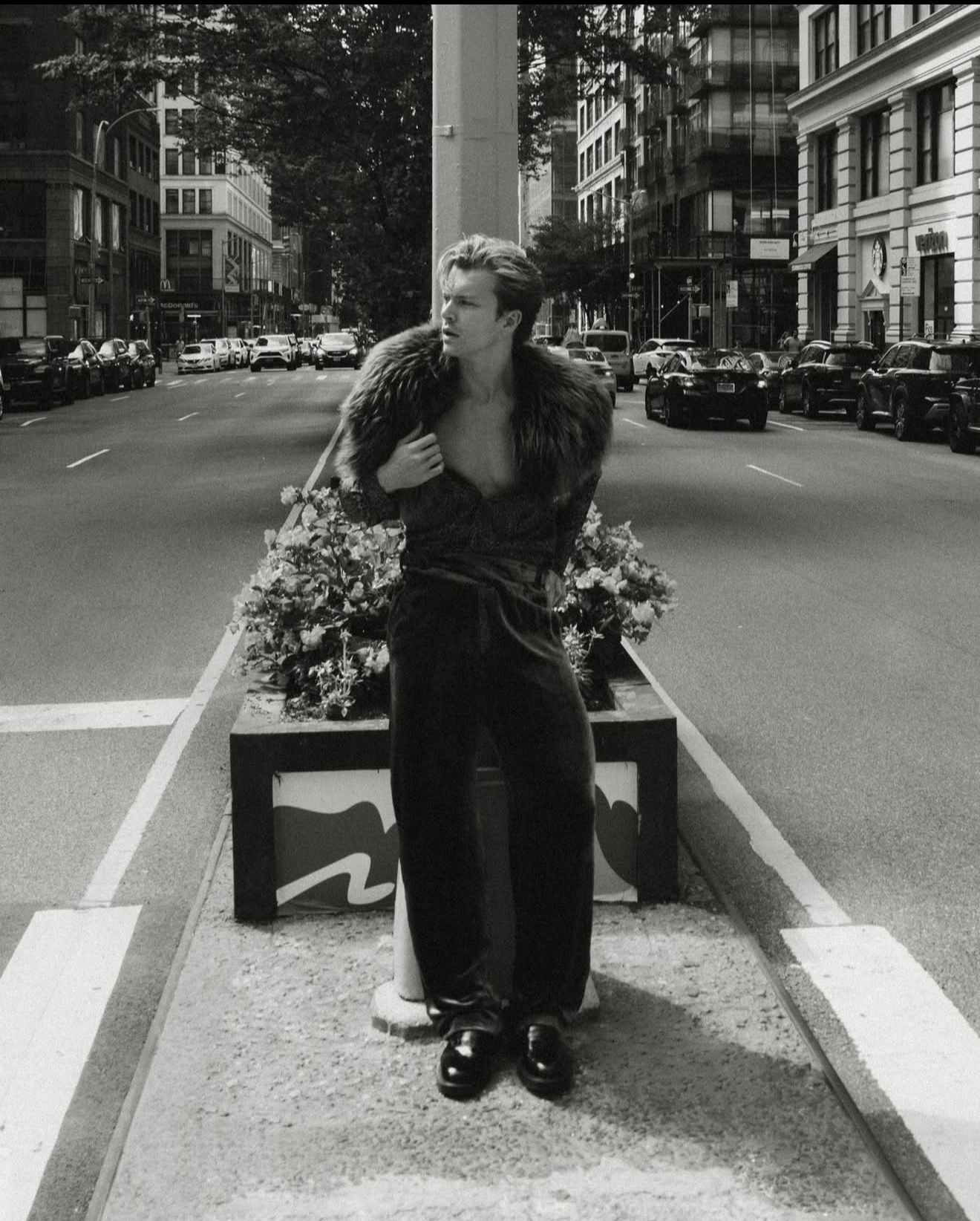 A black and white photo of a young man standing on a city street, posing in front of a flower bed, wearing a fur-collared coat and shiny shoes.