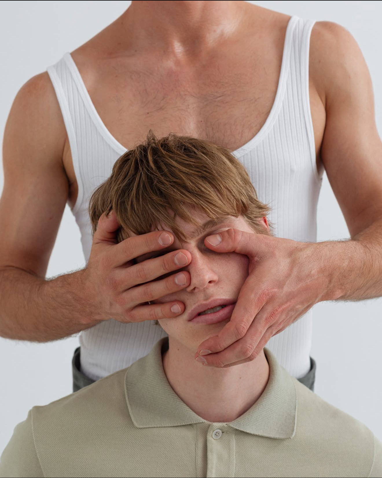 A man in a white tank top is holding a young boy's face, covering one eye with his hand and pushing the boy's face into his palm, creating an illusion of the boy's face being part of the man's chest.