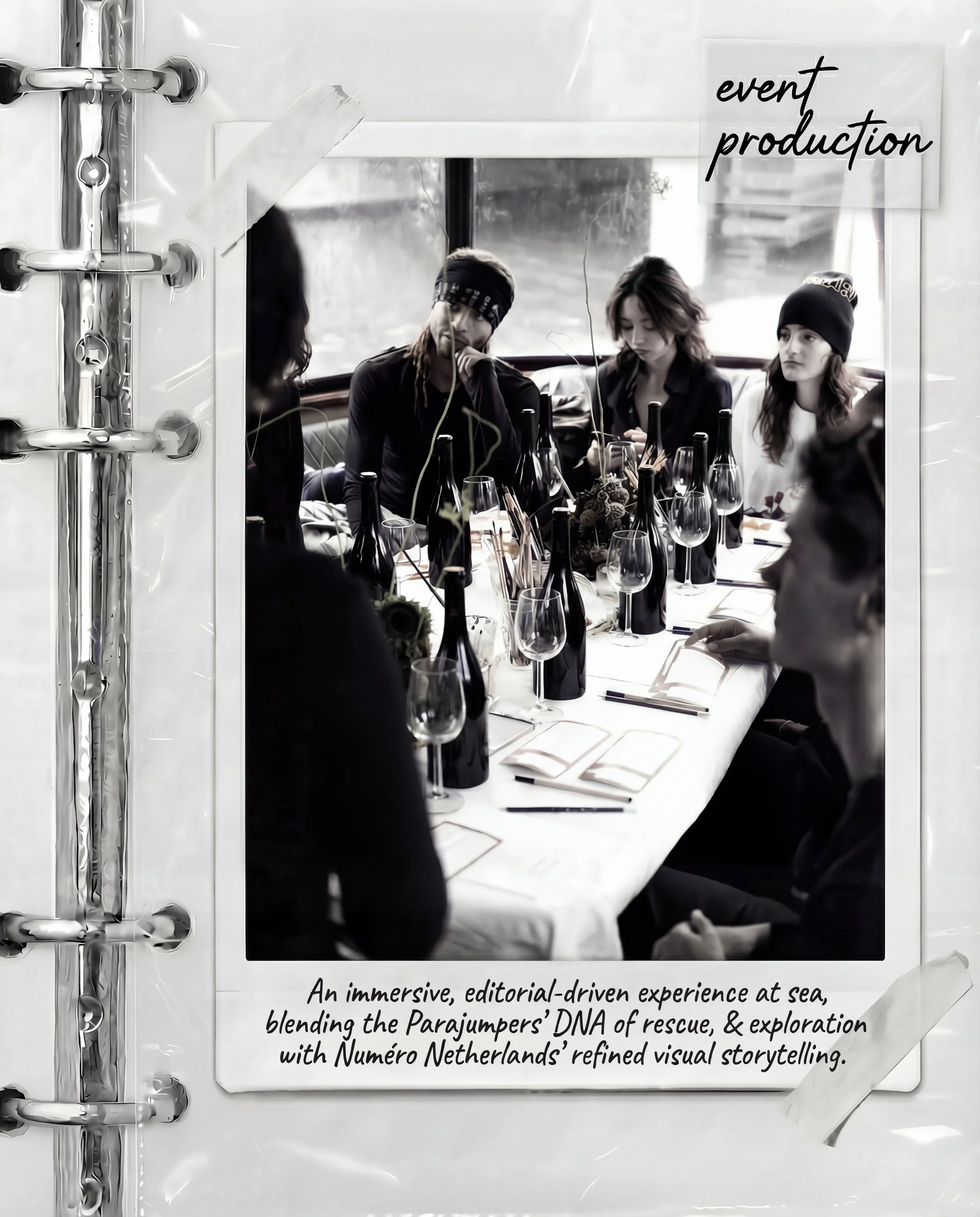 Black and white photograph of a group of people seated around a table with wine bottles and glasses, seen through a window with 'event production' written on a sign. The scene appears to be a meeting or gathering indoors.
