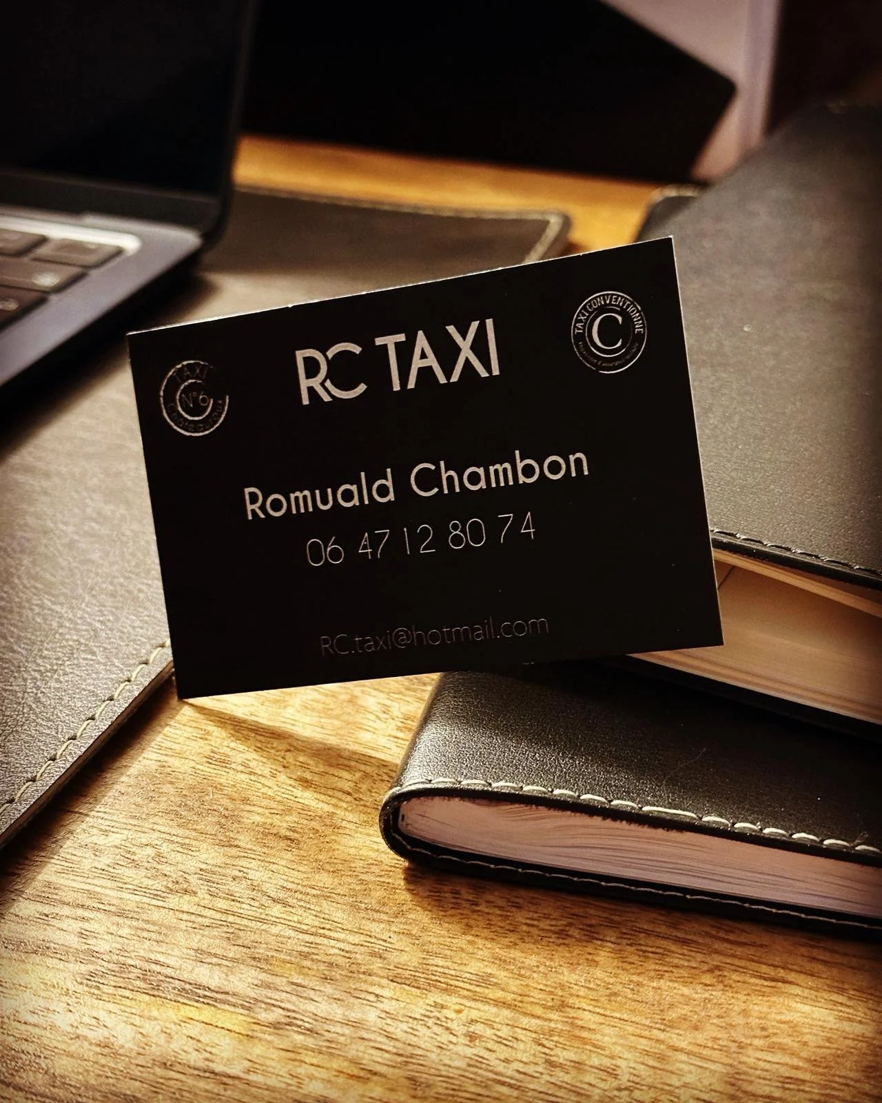 Carte de visite noir pour RC Taxi avec le nom Romuald Chambon, téléphone, adresse email, posée sur un carnet en cuir noir sur une table en bois.