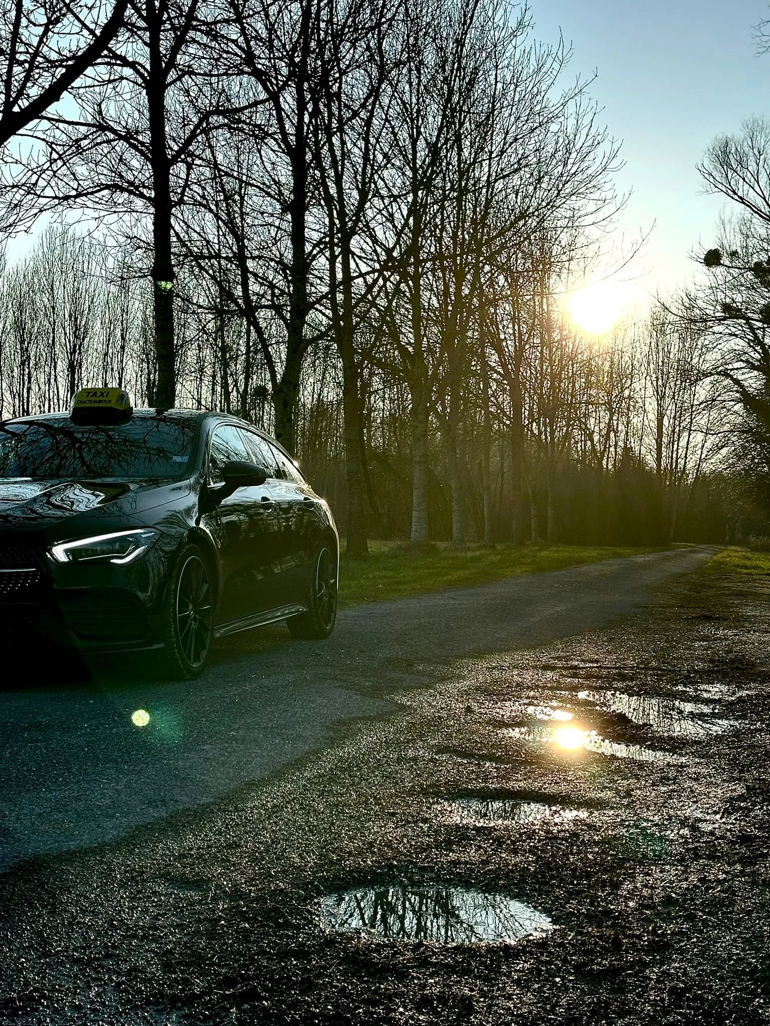 Voiture noire stationnée sur une route en forêt, avec des flaques d'eau réfléchissant le ciel, au coucher du soleil, arbres sans feuilles.