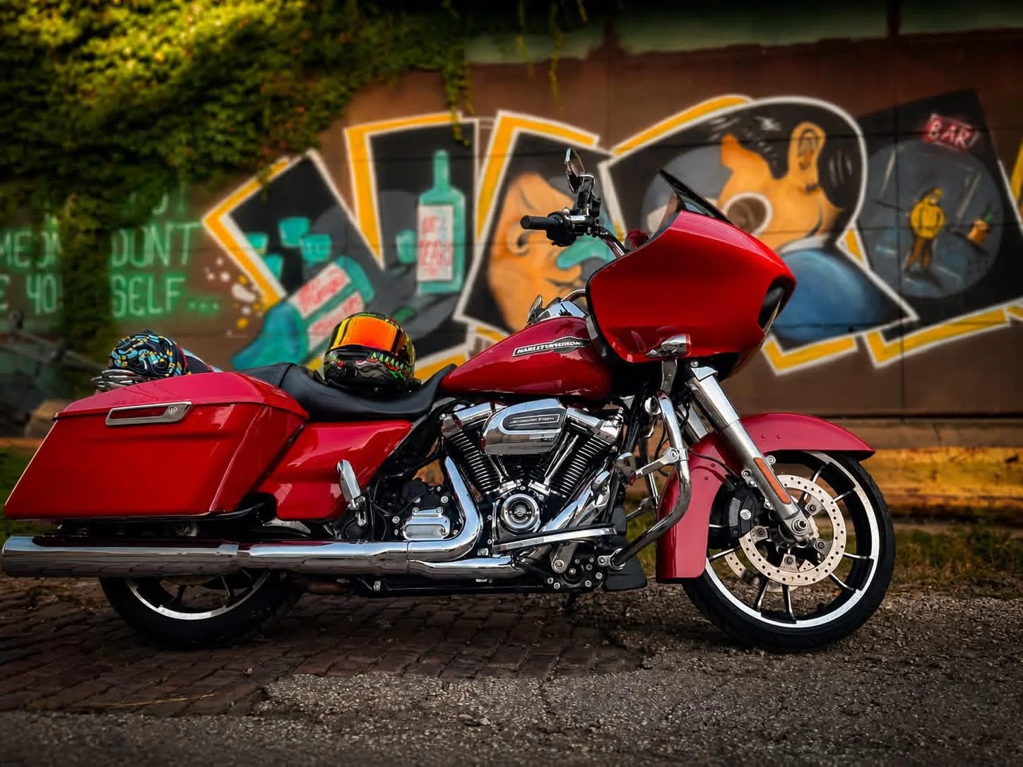 A red touring motorcycle parked on a cobblestone street, with two helmets on the seat and a colorful graffiti mural in the background.