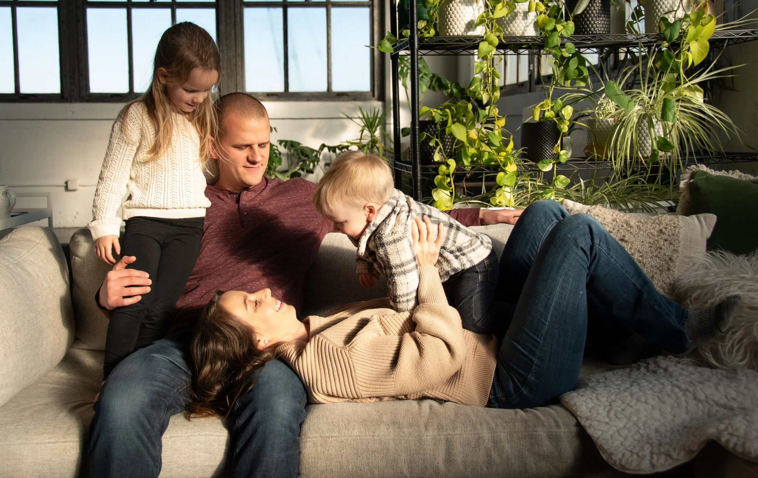 Family of four, two children and their parents, playing and relaxing on a beige sofa in a bright, plant-filled room.