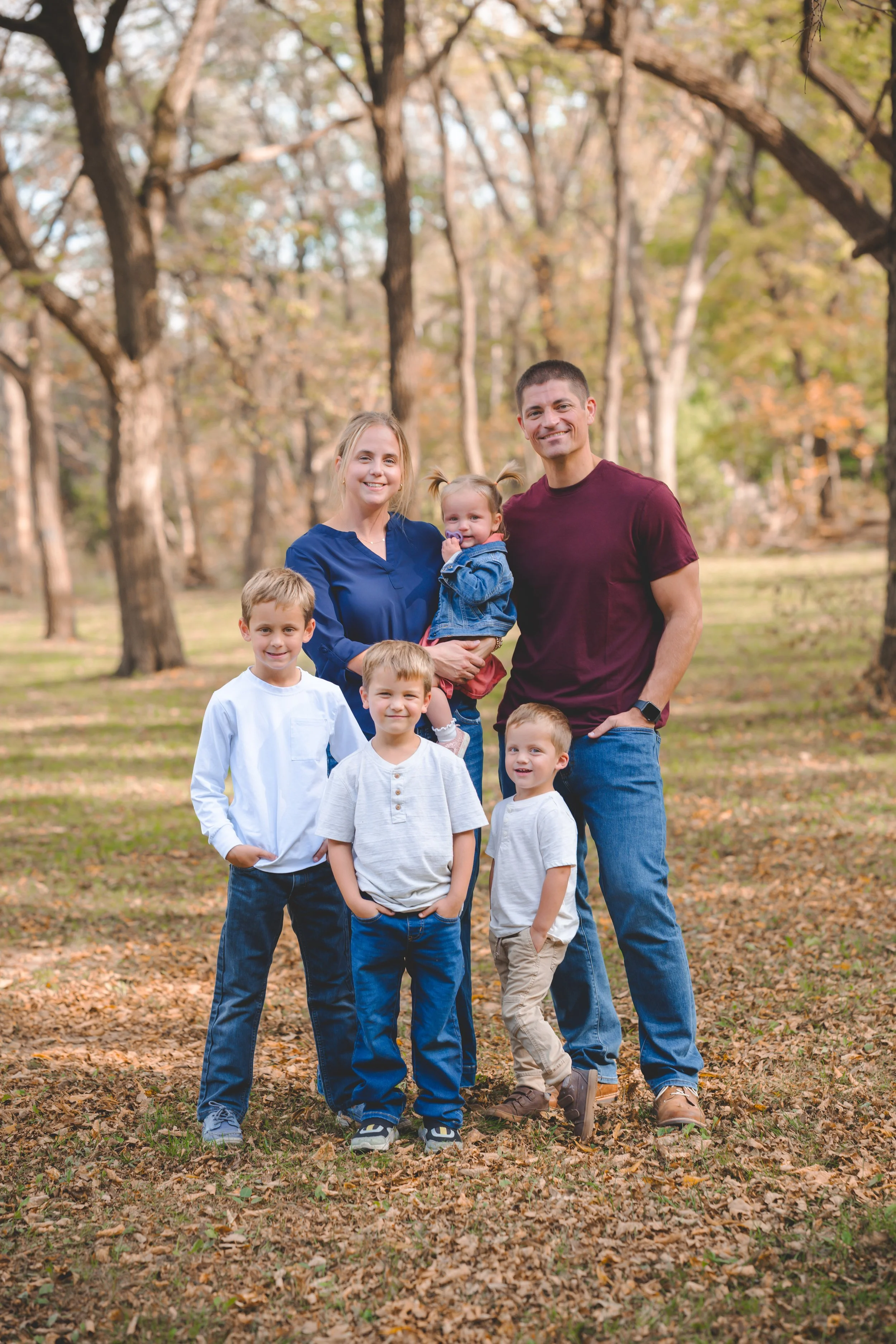 A family of seven outdoors in a park during autumn, with trees and fallen leaves in the background. The family includes two adults and five children, all smiling and dressed casually.