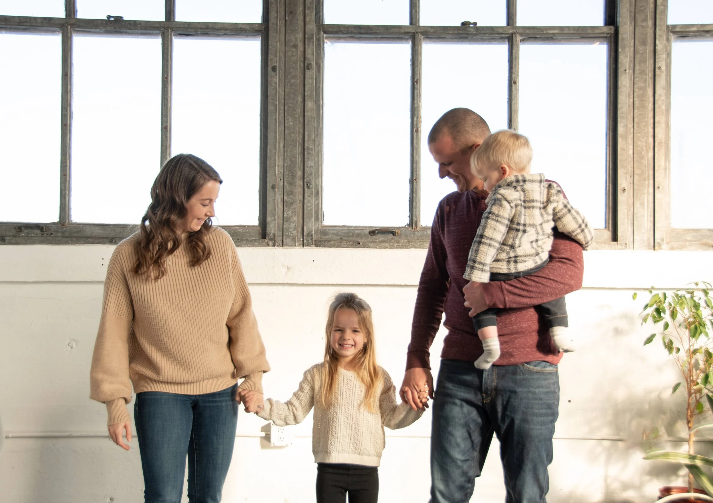 A family of four holding hands and smiling indoors near a large window with a white wall and a plant in the corner.