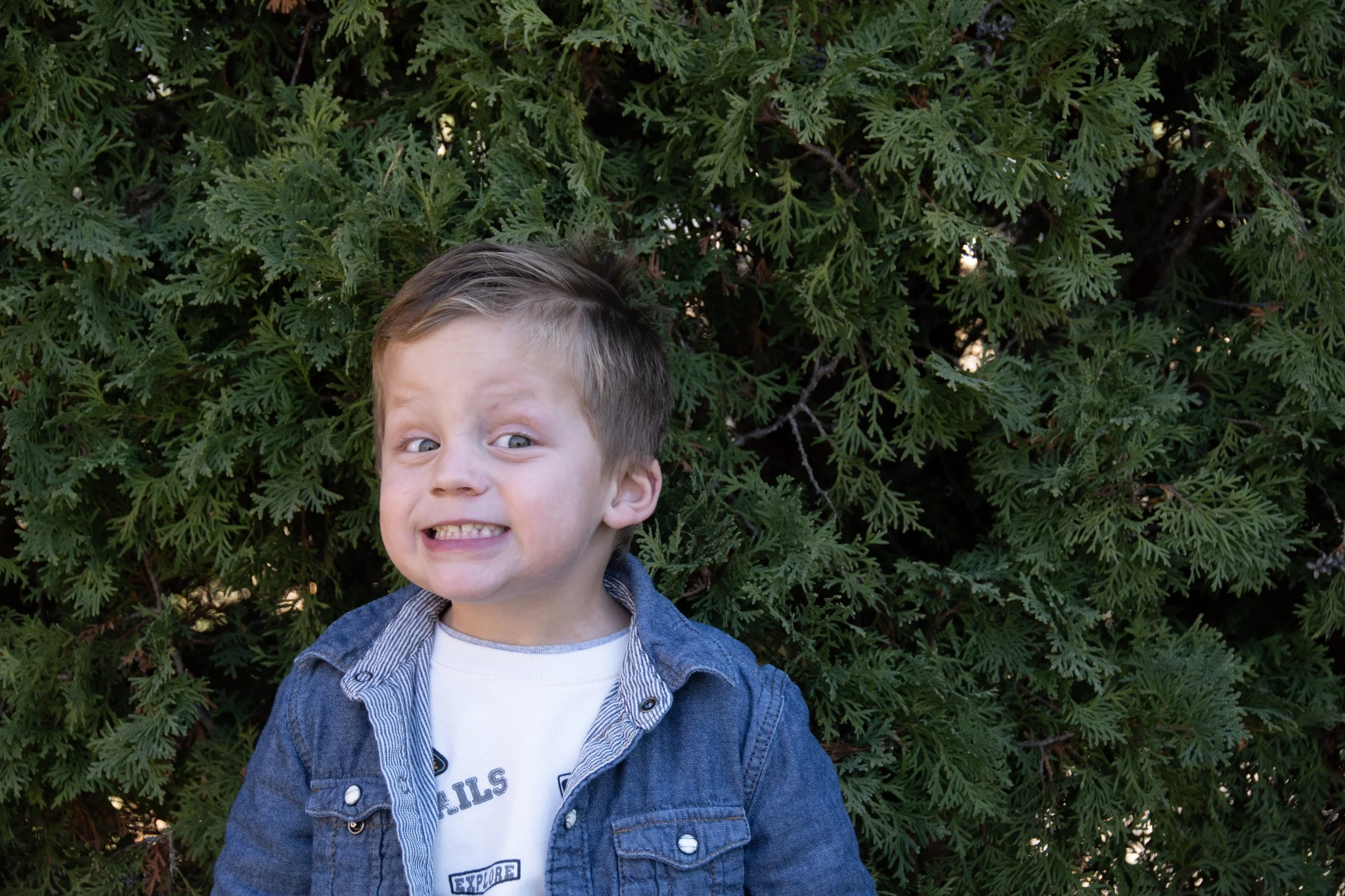 A young boy with a playful, scrunched-up facial expression, wearing a denim jacket over a white t-shirt, standing in front of a lush green bush.
