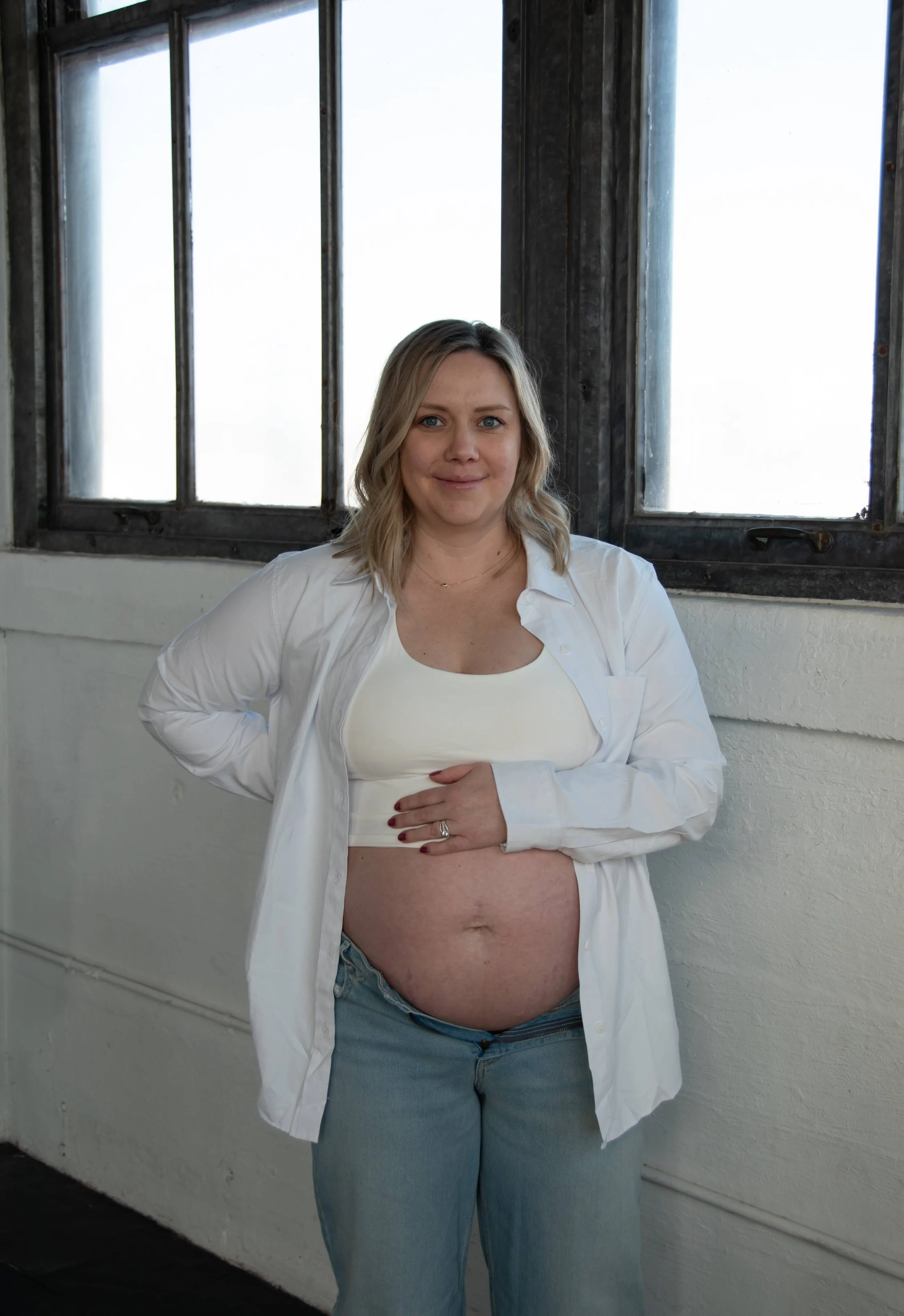 A pregnant woman with blonde hair, wearing a white tank top and an open white shirt, standing indoors near a window with black frames. She is smiling and has her hand on her belly.