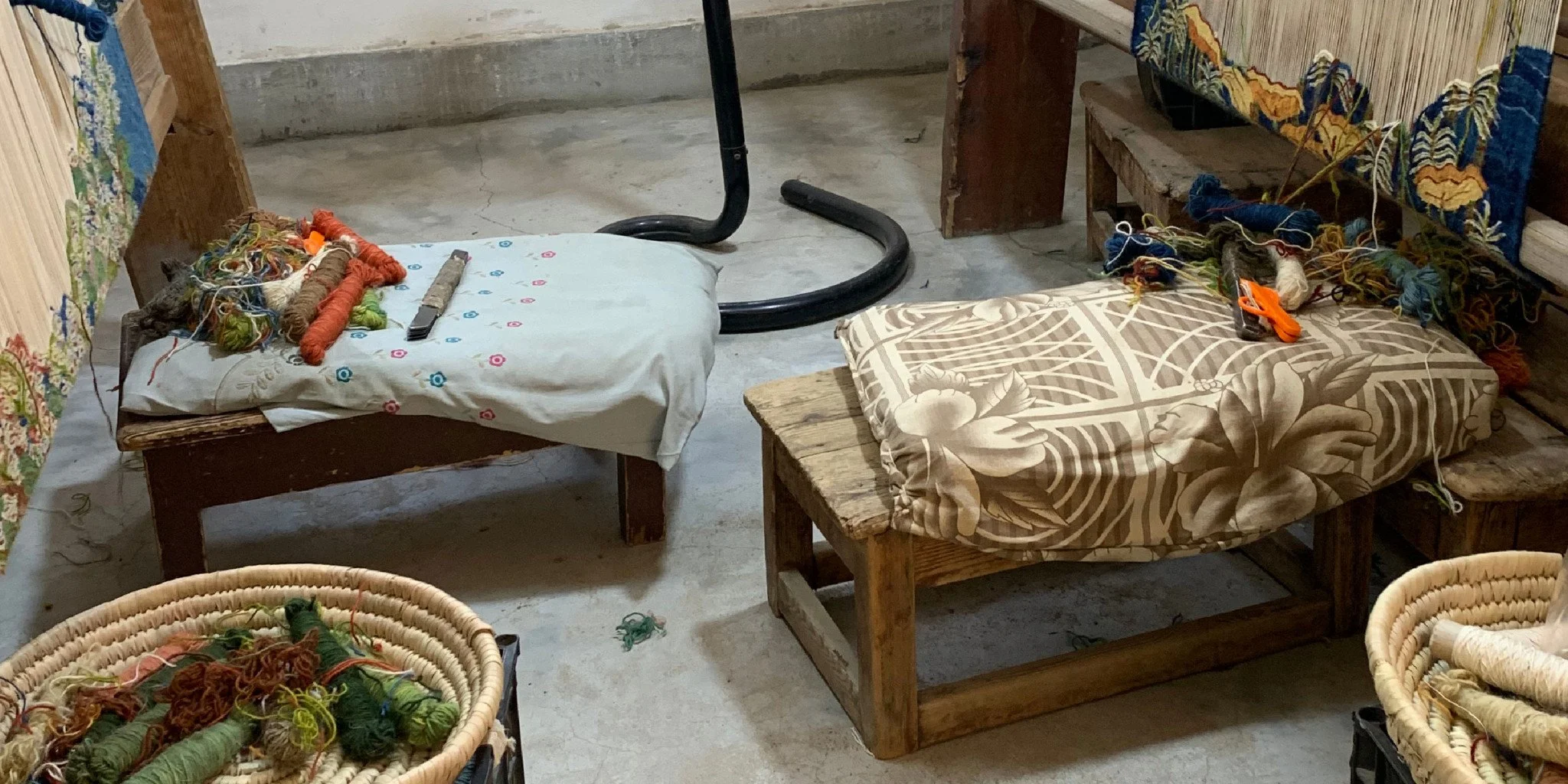 Two wooden beds with pillows, cushions, and yarn scattered on top, surrounded by baskets of yarn and a sewing tool, in a craft or workshop space.