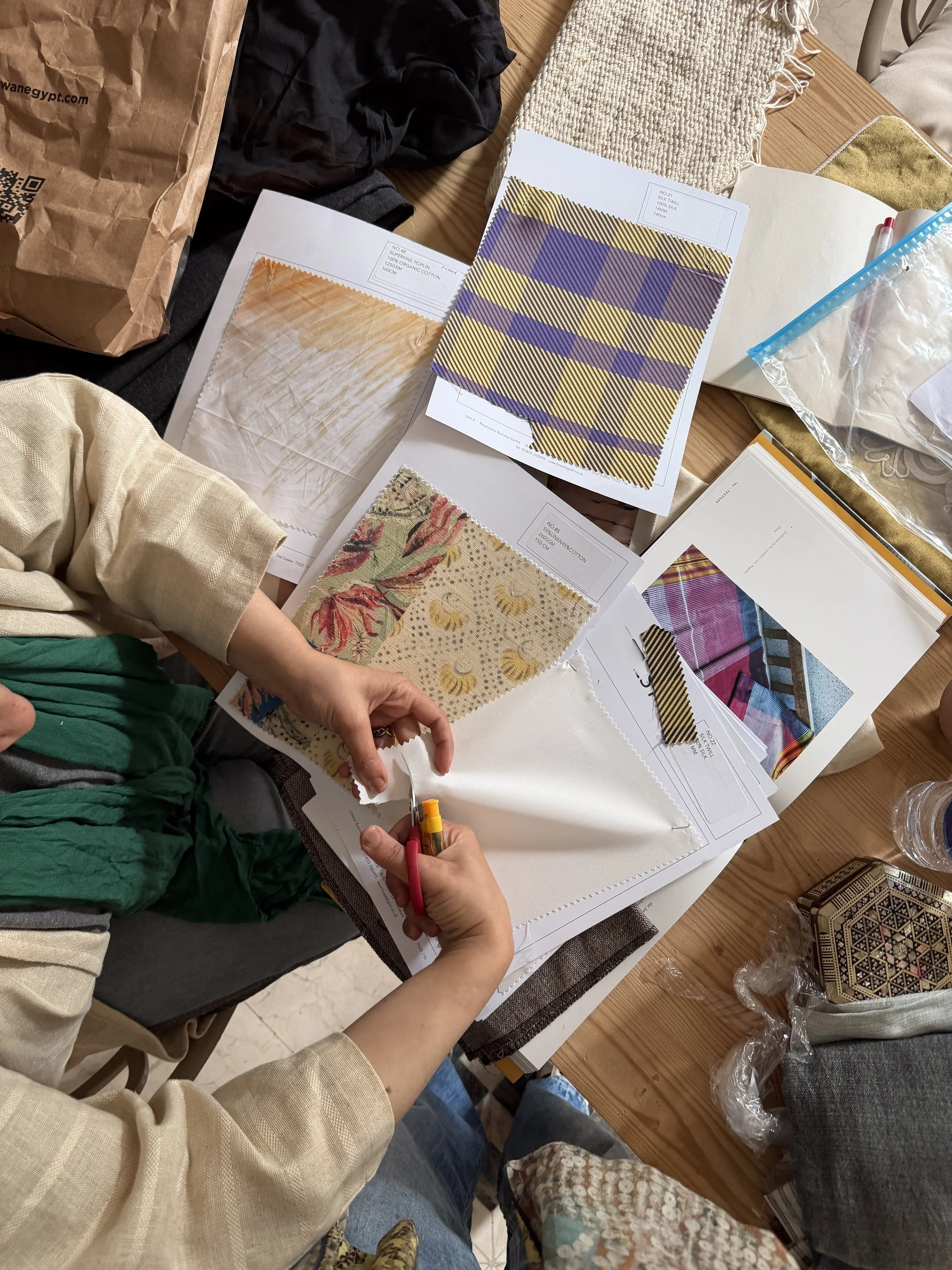 Person cutting fabric sample swatches with scissors on a cluttered table containing fabric swatches, design books, and sewing supplies.