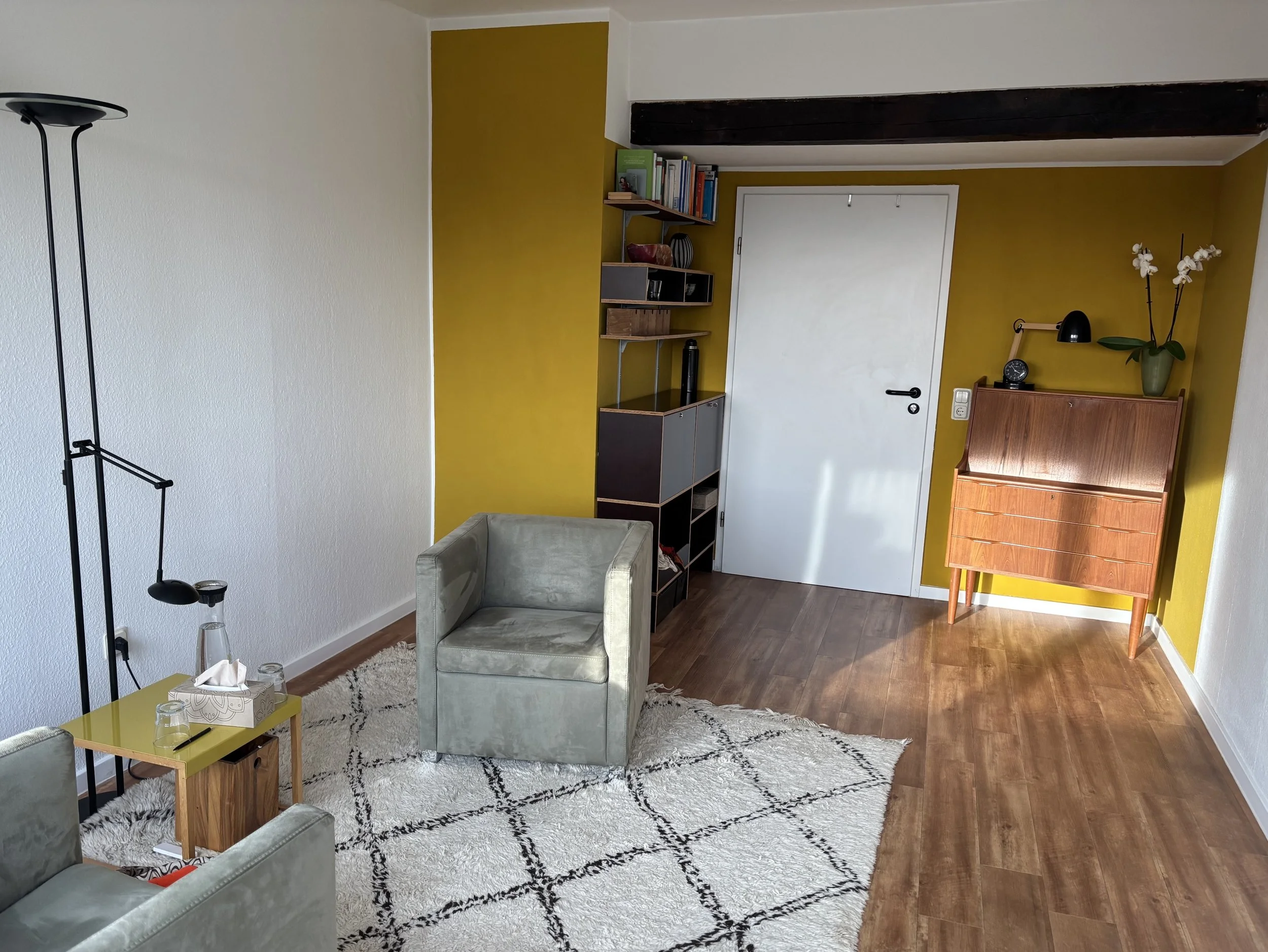 Wohnzimmer mit grauem Sessel, einem kleinen gelben Beistelltisch, einem weißen Teppich mit schwarzen Mustern, einer Stehlampe, einer Holzkommode, einer Wand mit dunkelgrünem Regal, einer weißen Tür und einer gelben Wand.