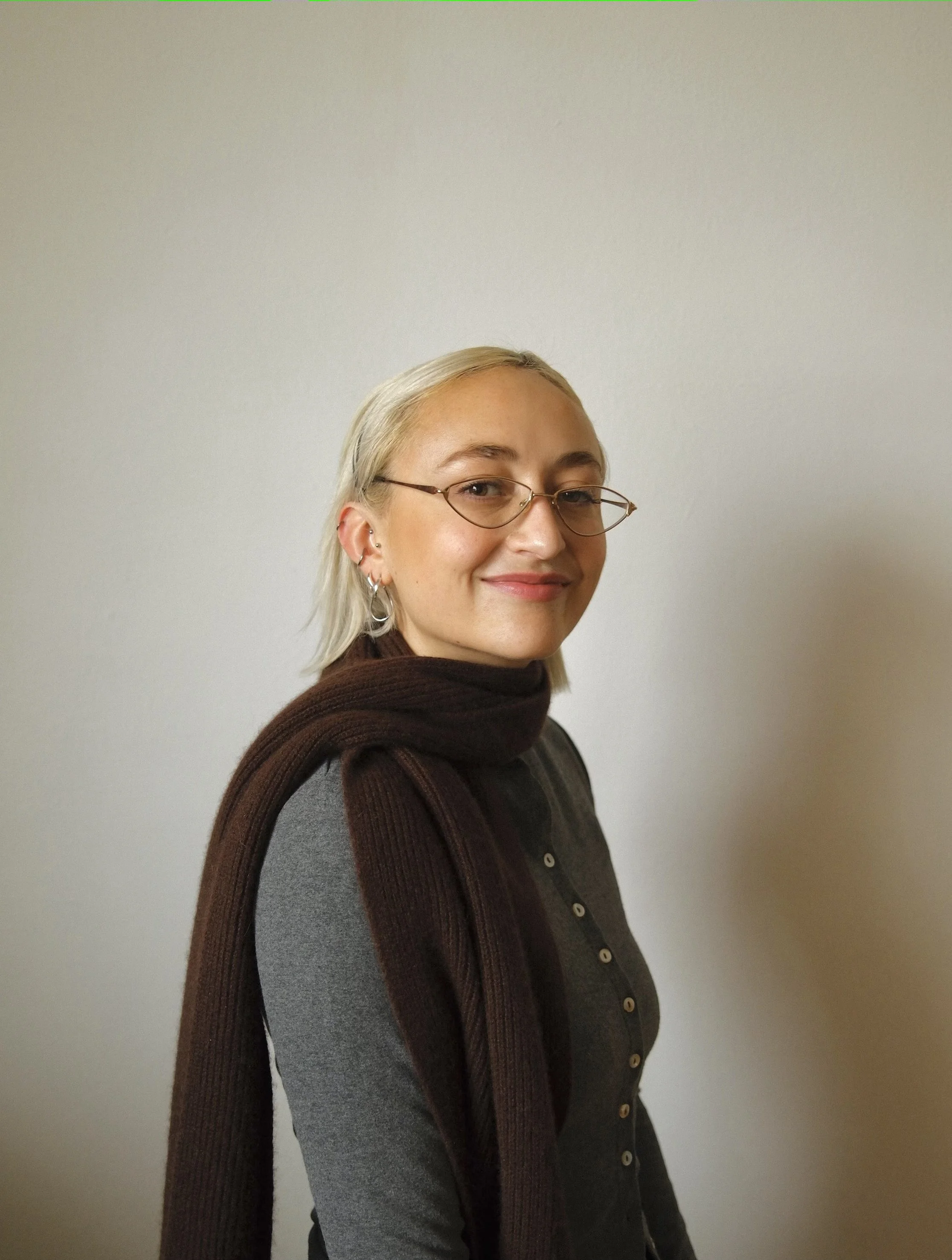 Porträt einer lachenden Frau mit blondem Haar, Brille, Ohrringen, grauem Oberteil und braunem Schal vor einer hellen Wand.