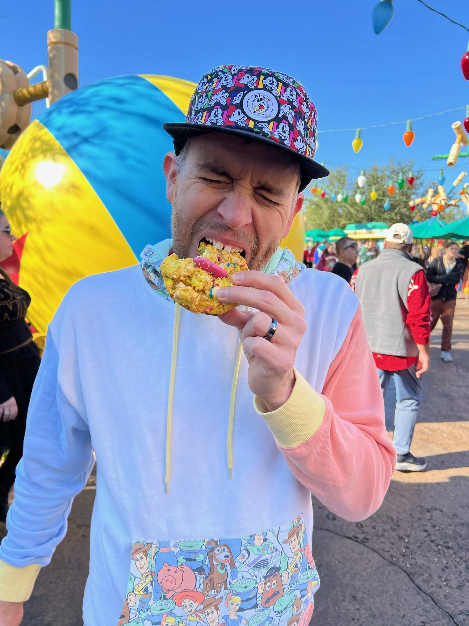 A man eating a cookie at a theme park with colorful decorations and large inflatable character in the background.