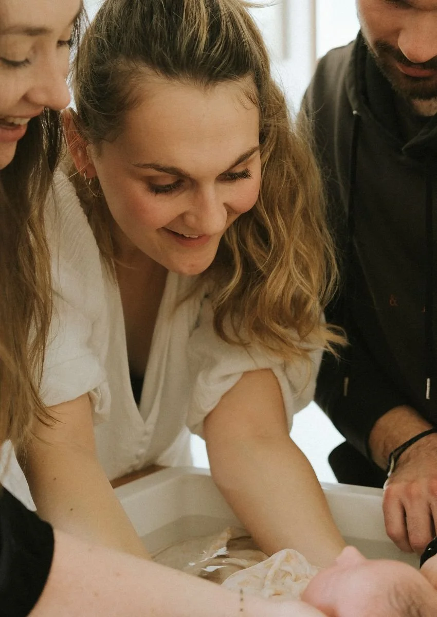 Groupe de trois personnes souriantes, avec deux femmes et un homme, qui regardent attentivement un bébé lors du bain ou d'une activité de soins.