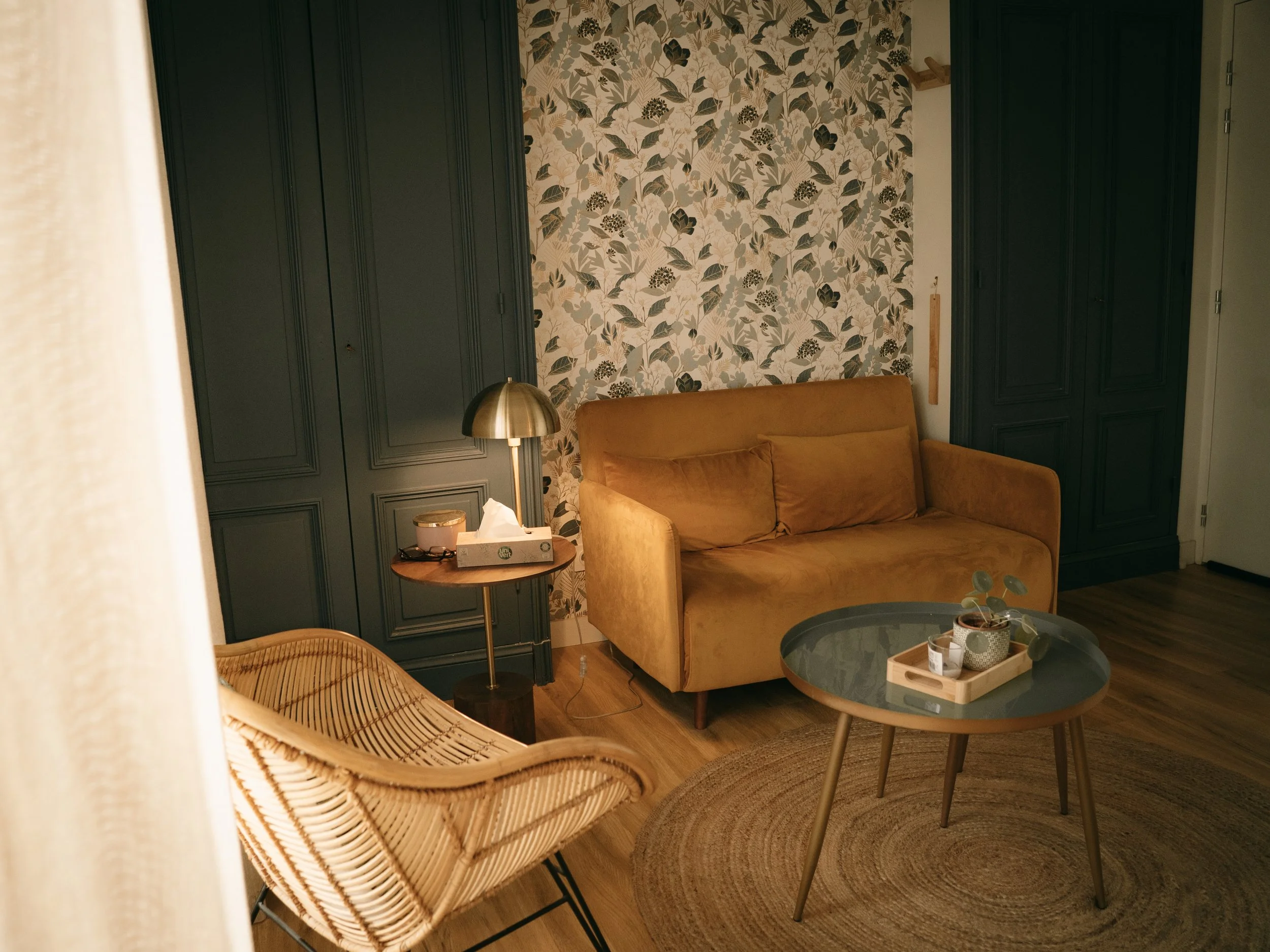 Salon avec un canapé beige, une table en bois avec un plateau en verre surmonté de plantes en pot, un fauteuil en rotin, un lampadaire doré, un tapis rond en jute, et un mur avec du papier peint à motifs végétaux, entouré de placards en bois foncé.