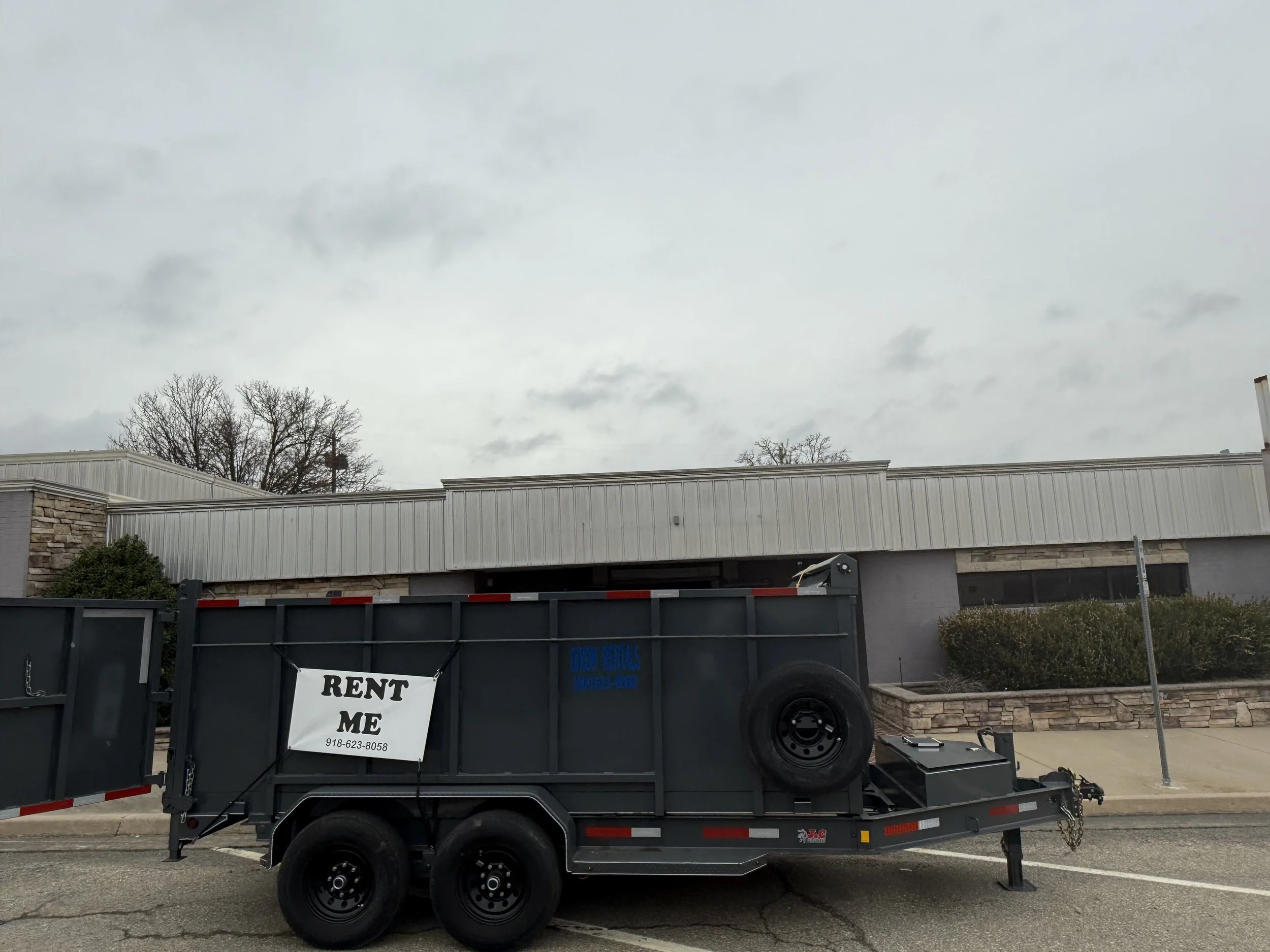A black trailer parked on a street with a sign that says 'RENT ME' and a phone number, in front of a gray building with bushes and leafless trees under a cloudy sky.