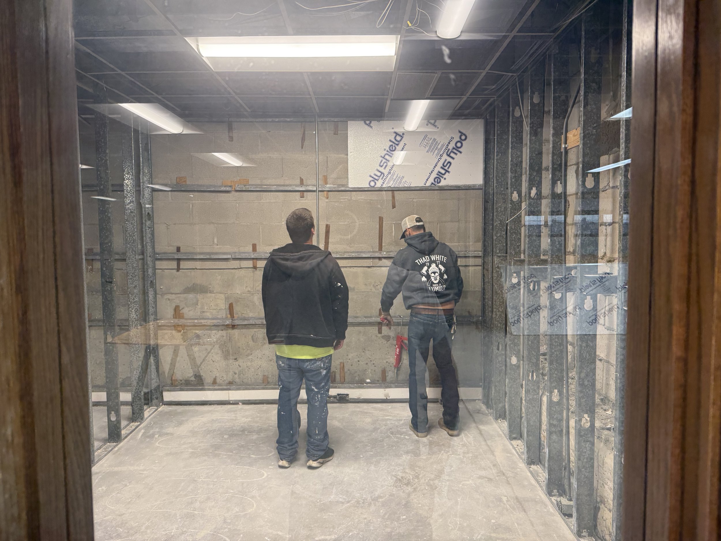 Two construction workers inside a small, unfinished room with metal framing, concrete walls, and a ceiling with fluorescent lights, viewed through a glass window.