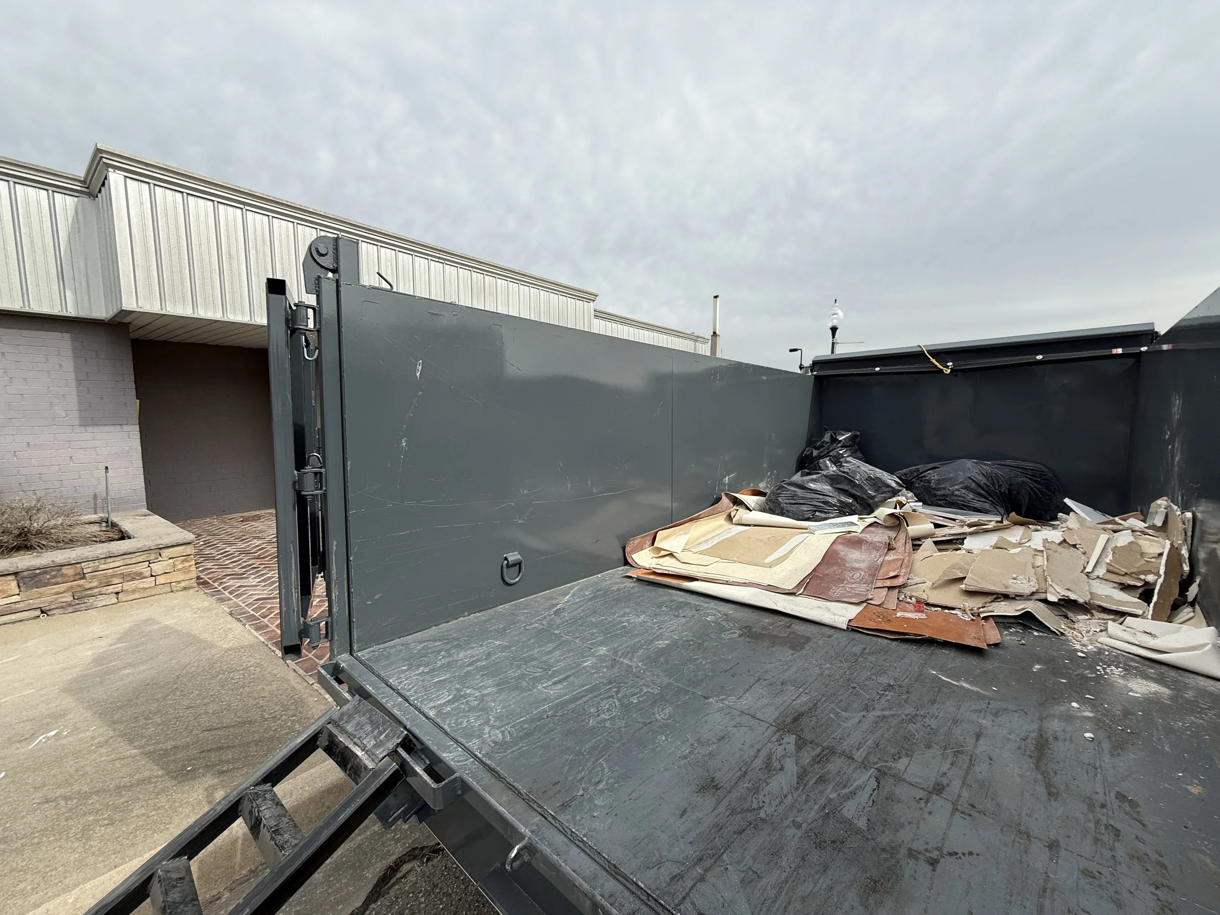 Back of a grey dump truck filled with black garbage bags, cardboard, and debris, parked near a building with a brick pathway and a small front yard.