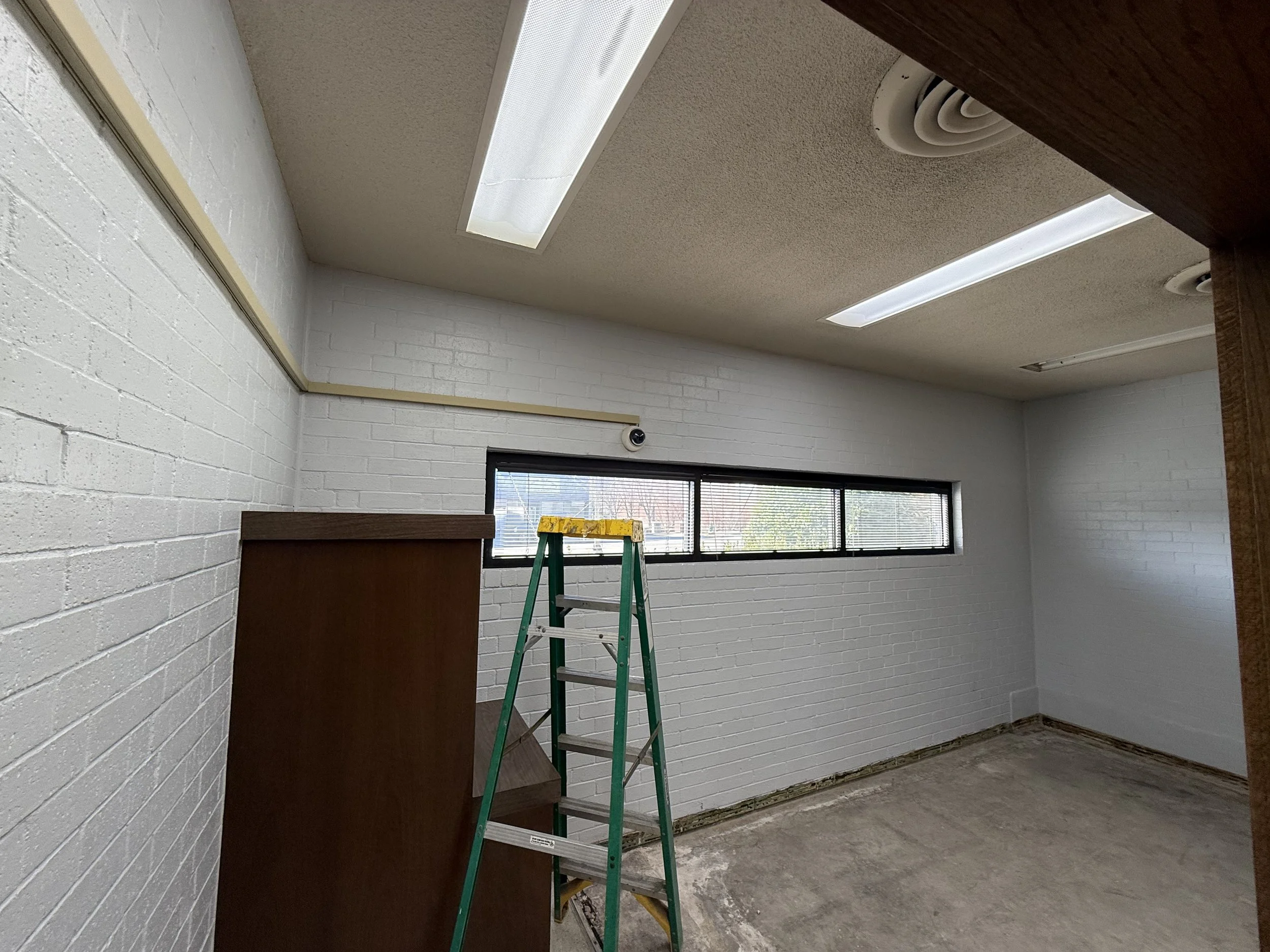 Empty room with white brick walls, rectangular window, fluorescent ceiling lights, green ladder, and a wooden cabinet.