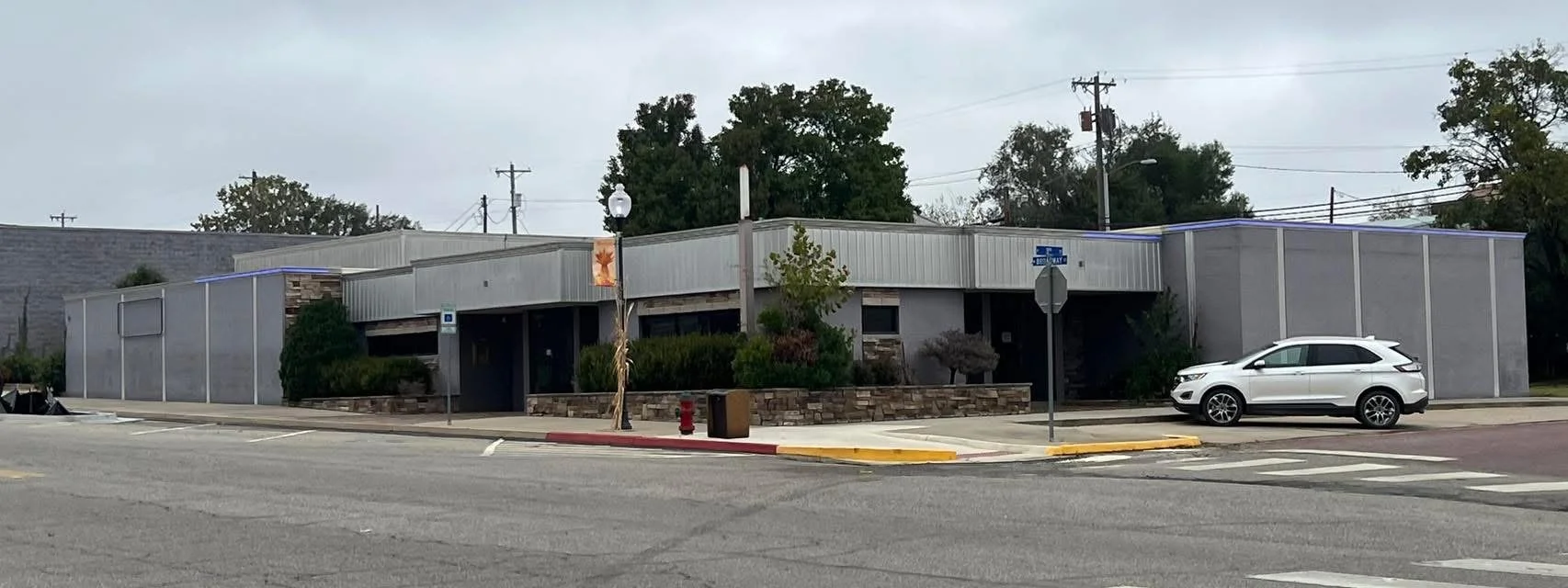 Street view of a commercial building with a stone and metal exterior, parked white SUV, crosswalk, street signs, and trees in the background.