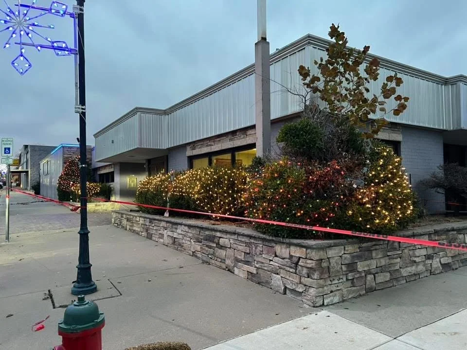 Street view of a commercial building decorated with Christmas lights, with bushes wrapped in string lights, and a sidewalk with a fire hydrant and a streetlamp, on a cloudy evening.