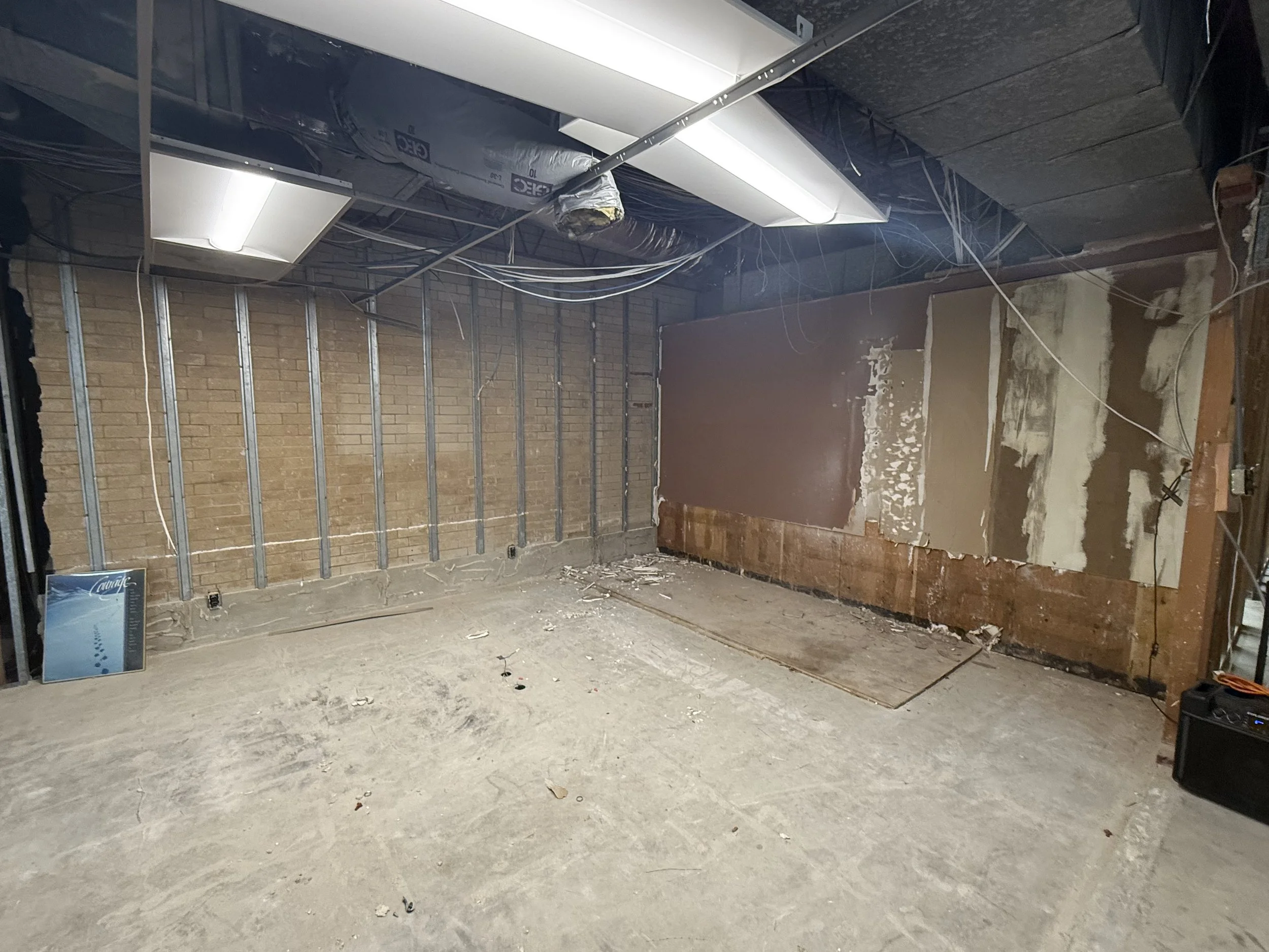 Unfinished room under renovation with exposed brick wall, electrical wiring, drywall in progress, and fluorescent ceiling lights.