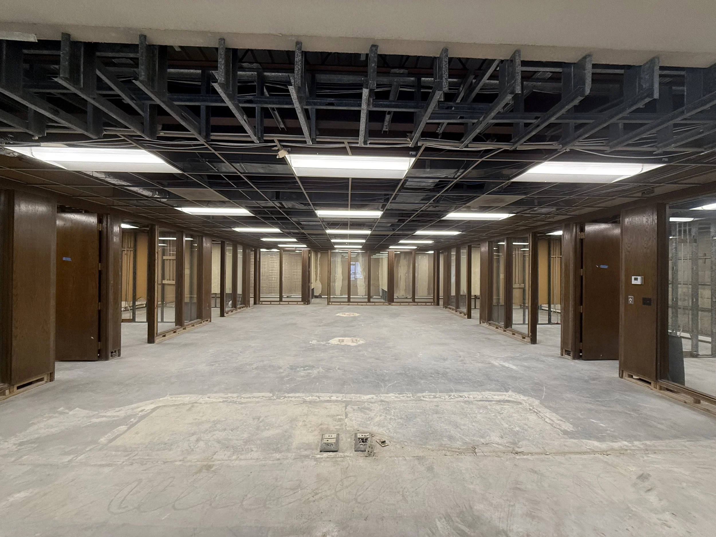 Empty office space under construction with exposed ceiling framework, wooden support beams, and glass partitions lining the walls.