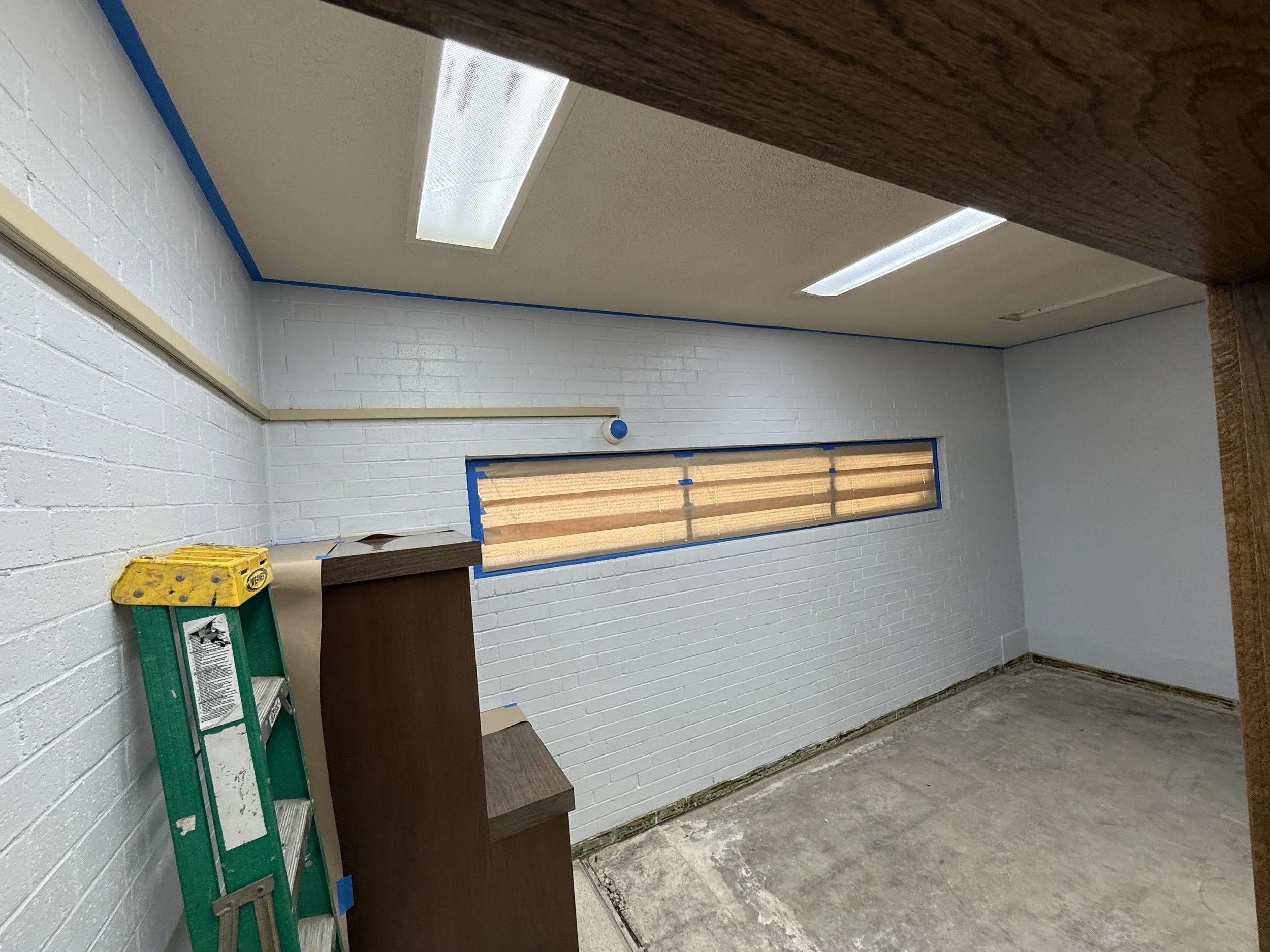 Room under renovation with white painted brick walls, a long horizontal window with wood blinds, and a concrete floor. Construction supplies include a green step ladder and a wooden cabinet.
