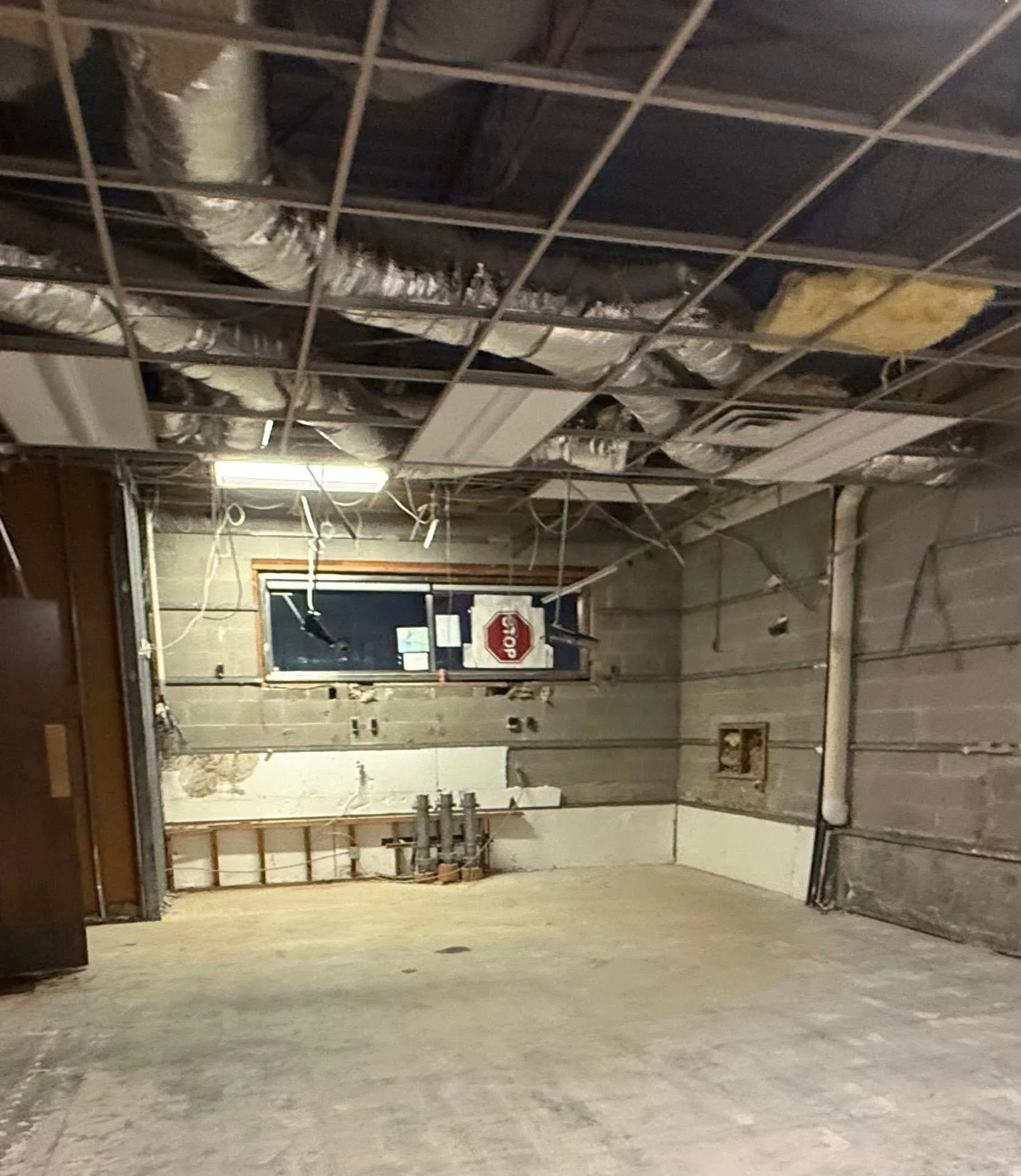 Empty garage with exposed ceiling ducts, pipes, and wires, unfinished walls, concrete floor, and a window with a stop sign outside.