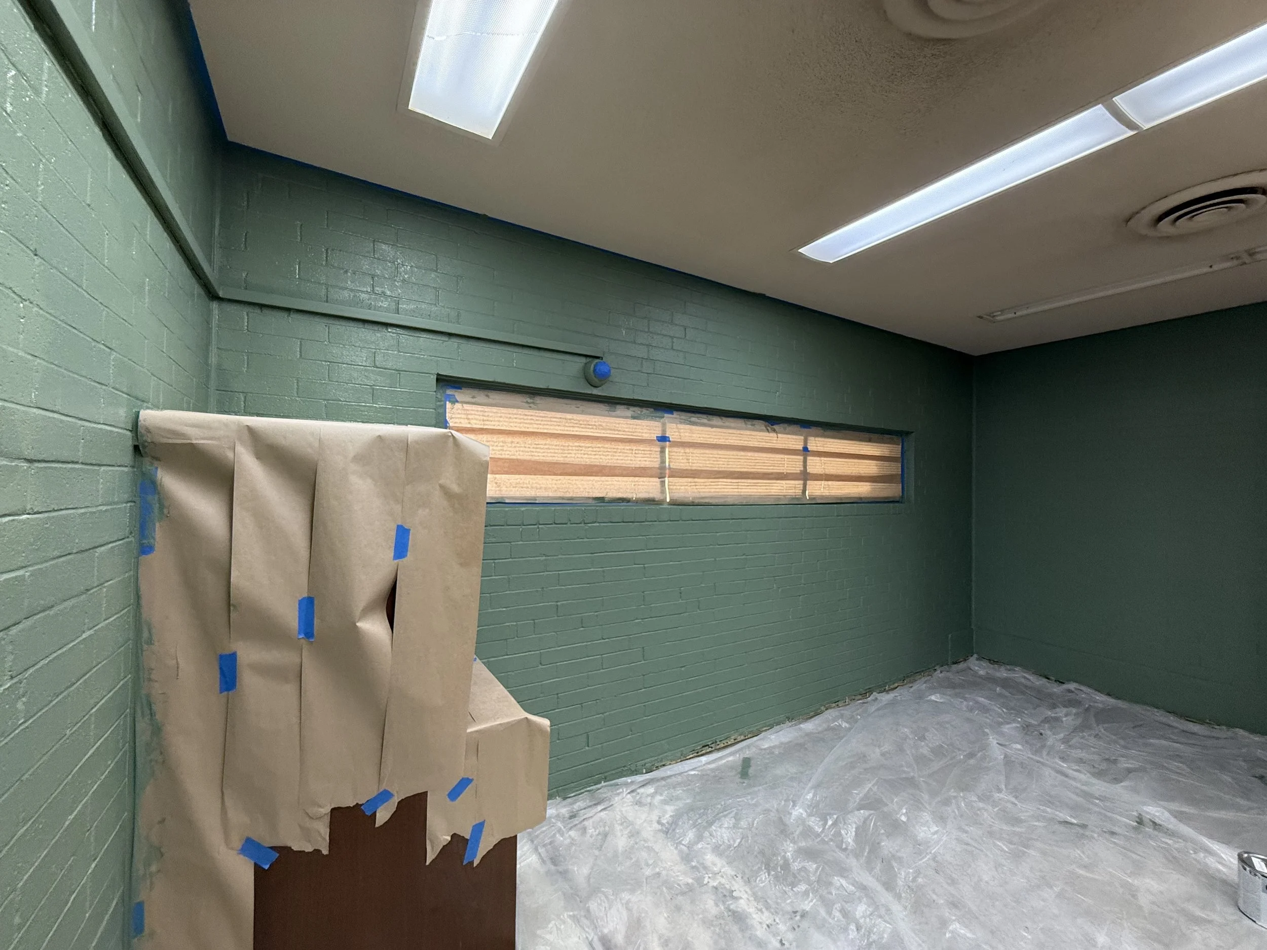 Empty room with green painted brick walls, a rectangular window covered with wooden slats, plastic sheets on the floor, and furniture wrapped in brown paper and taped off. Ceiling has fluorescent lights and a vent.