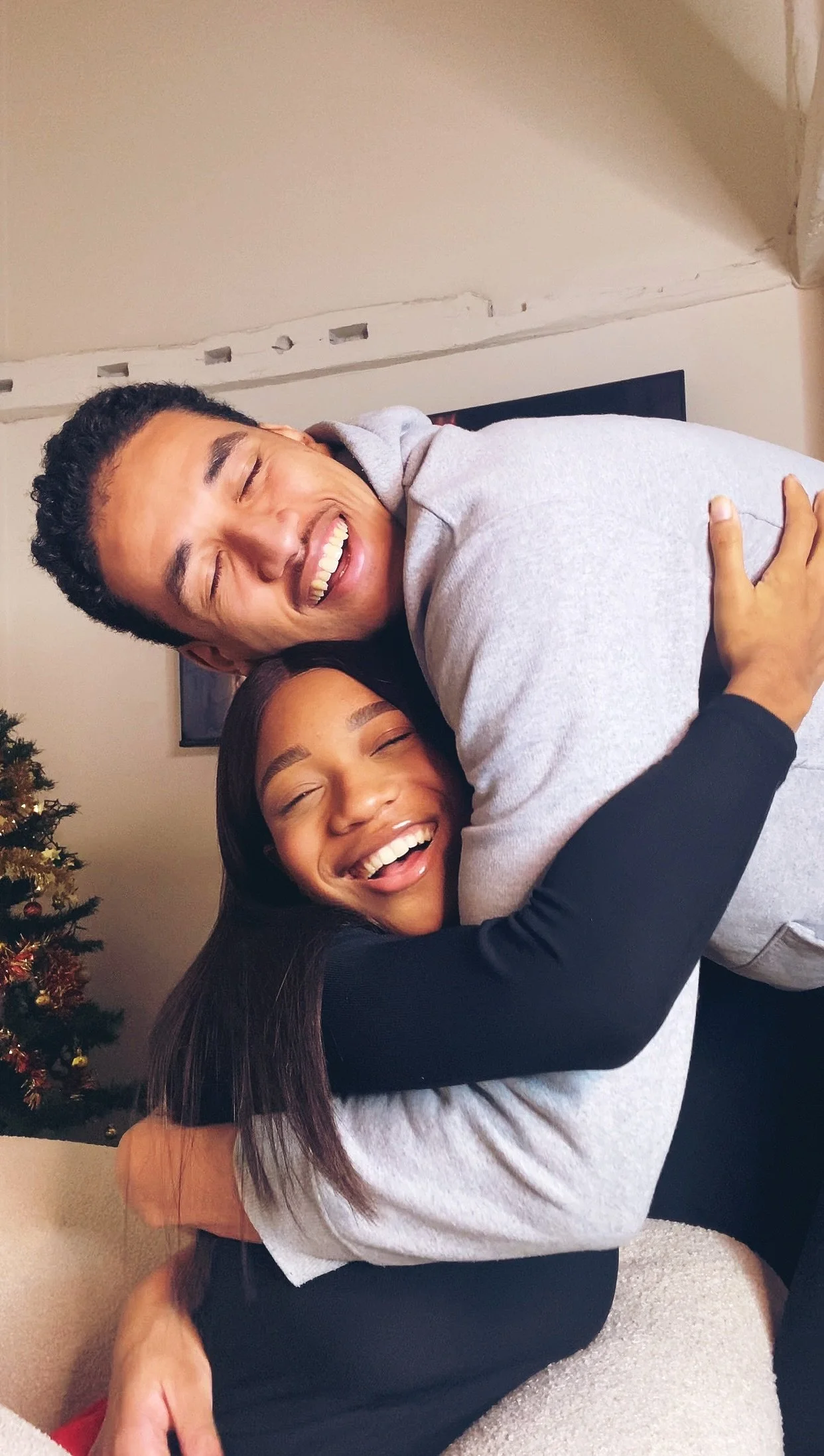 Two people hugging and smiling in a cozy room with a Christmas tree in the corner.