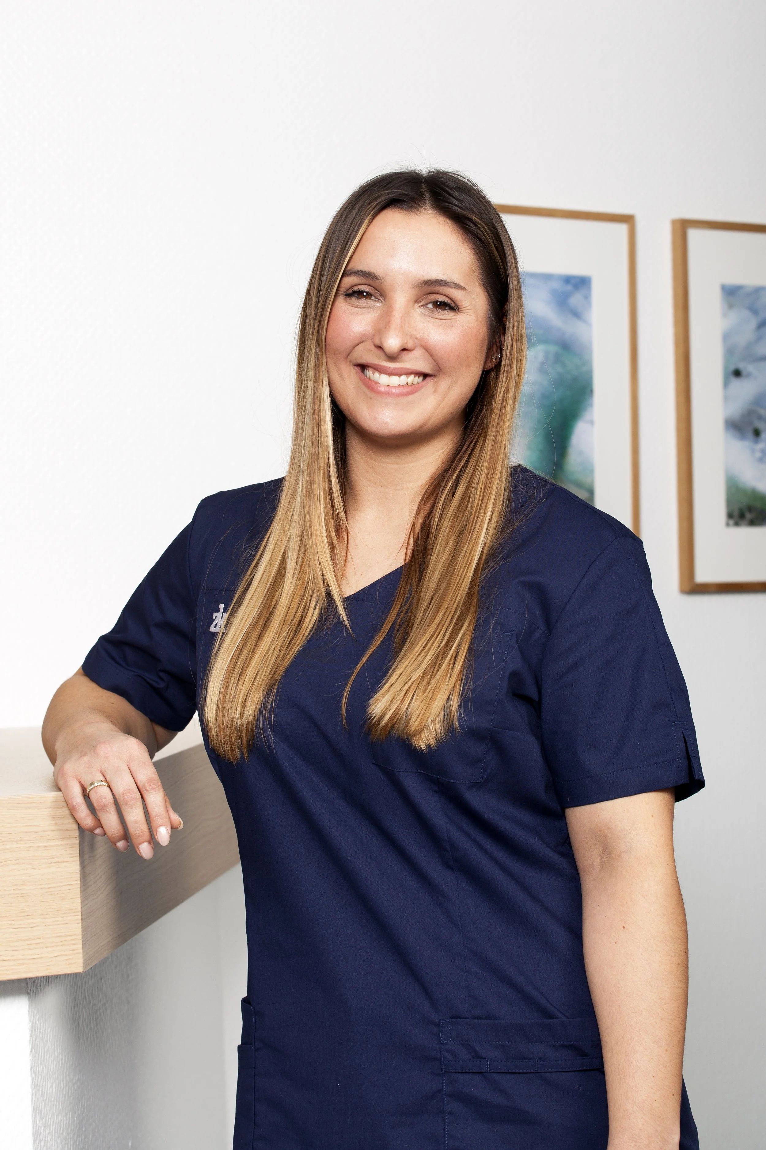 Tanée Bou, M.Sc. Kieferorthopädie in blauer Berufsbekleidung vor weißer Wand mit Bildern.