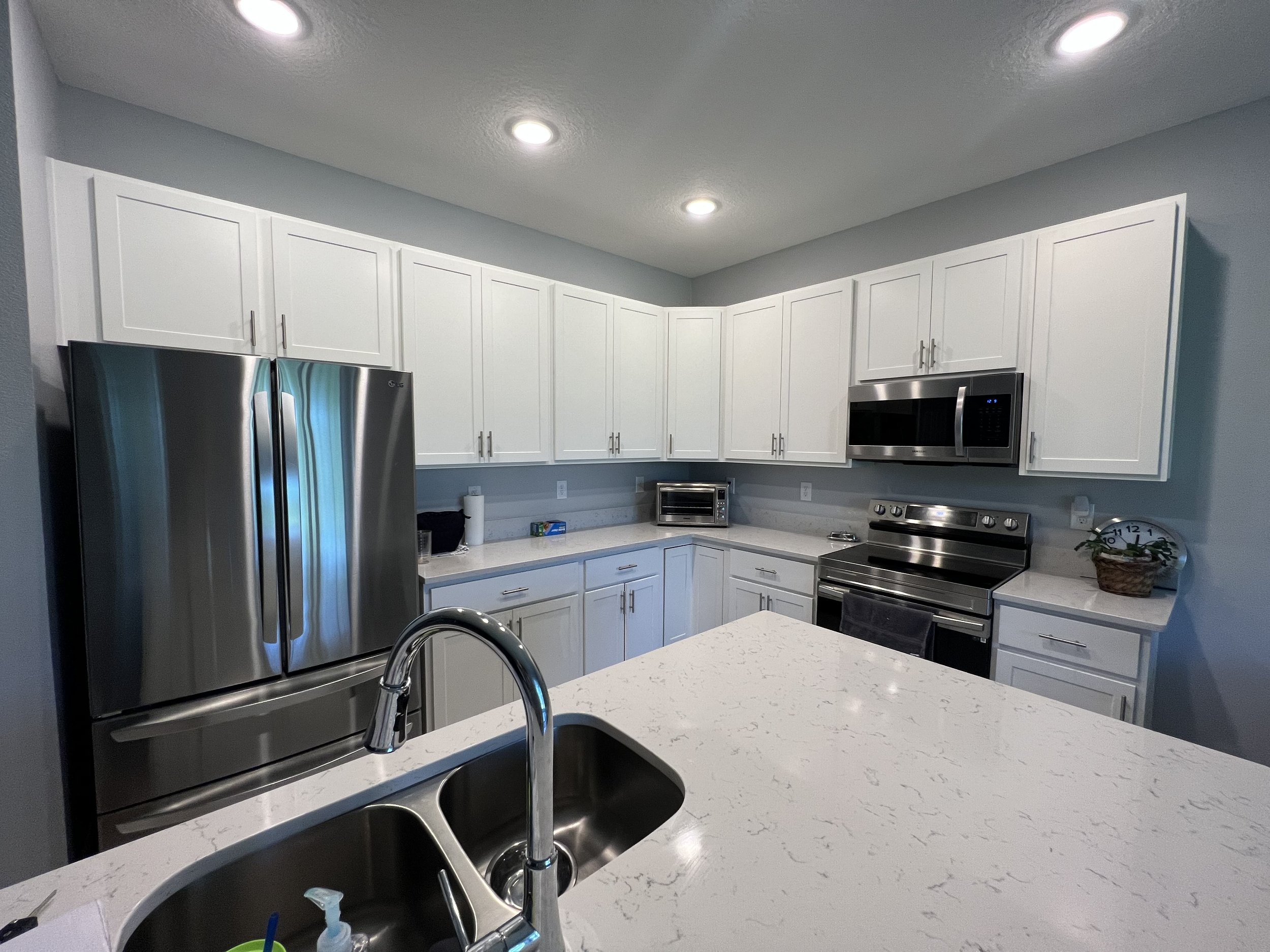 A modern kitchen with white cabinets, stainless steel refrigerator, microwave, oven, and a white countertop with a sink and faucet.