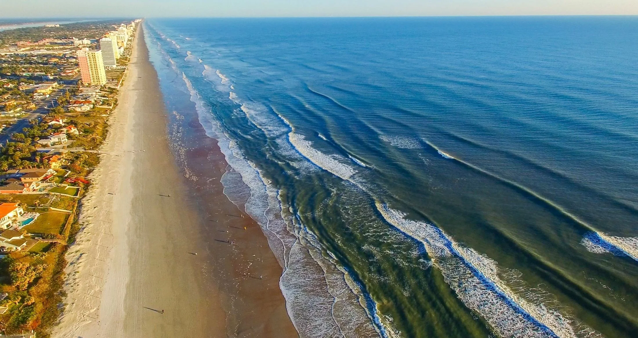 Aerial view of a coastal city of Daytona Beach Shores with high-rise buildings along a sandy beach and blue ocean waves.