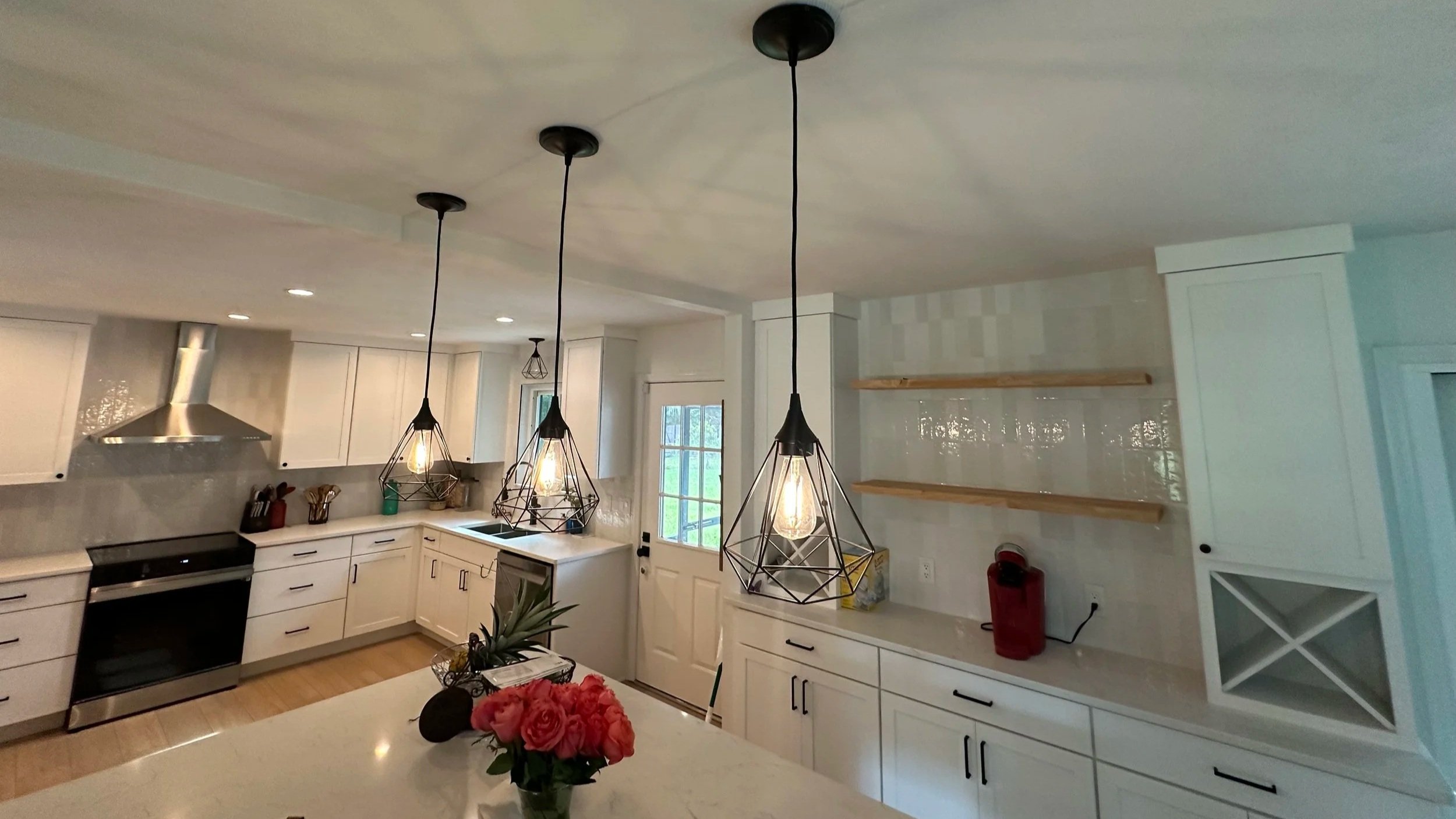 Modern kitchen with white cabinets, black hardware, a black oven, and industrial-style pendant lights hanging from the ceiling. There are open wooden shelves on the right wall and a window in the background. A vase with pink flowers is on the countertop.