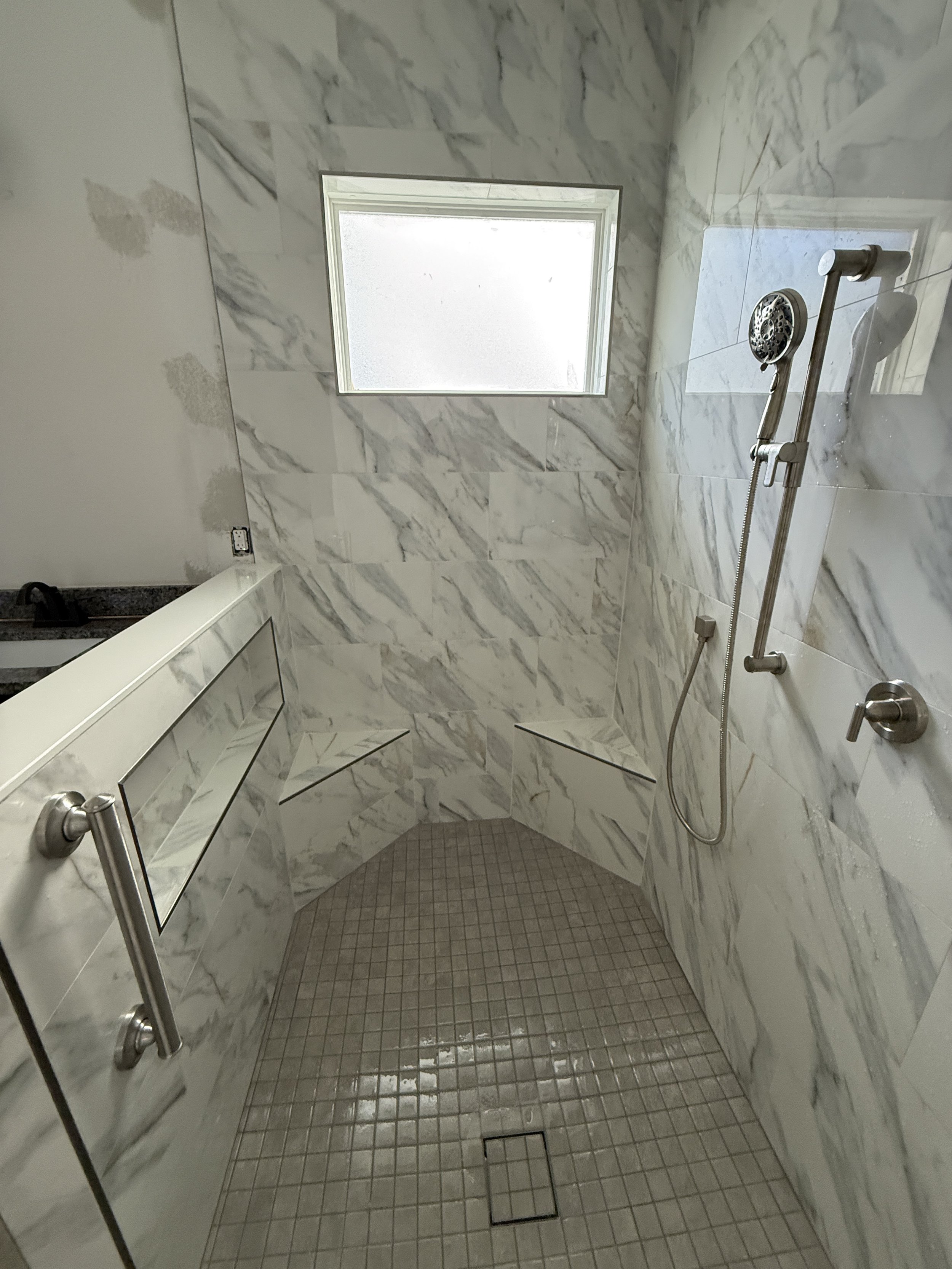 A custom built ADA compliant walk-in shower with tiled walls, a custom glass piece, a handheld showerhead with wal integration, two shower bench seating. and grab bars. Completed this in 2025 in Port Orange Florida. 